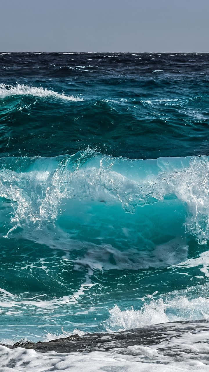 極致海洋浪花 iPhone 壁紙 — 在晴朗的天空下，一道強勁的藍綠色海浪拍打著深色岩石。