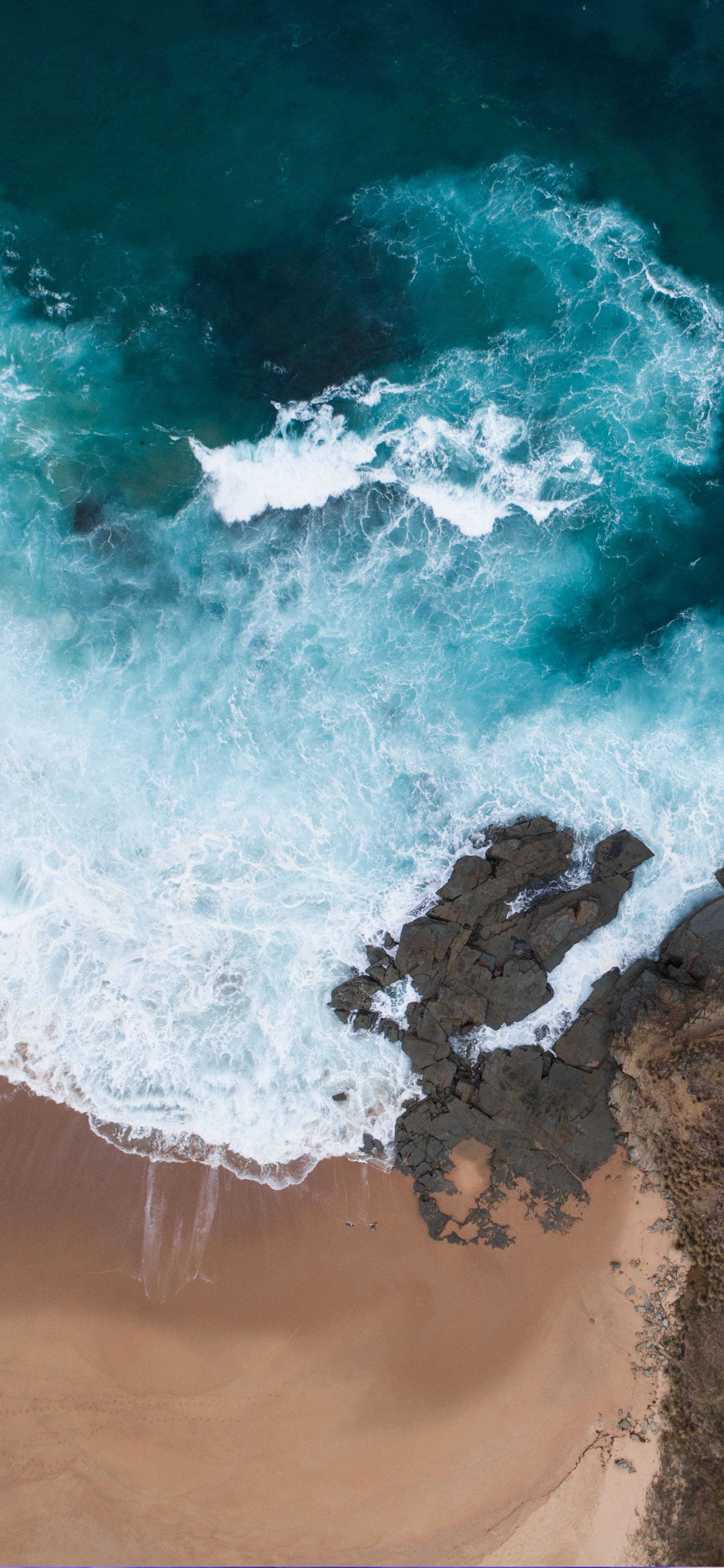 iPhone XS 海洋鳥瞰風景 壁紙 — 從高空俯瞰，碧綠色的海浪拍打著深色岩石，旁邊是金色沙灘。