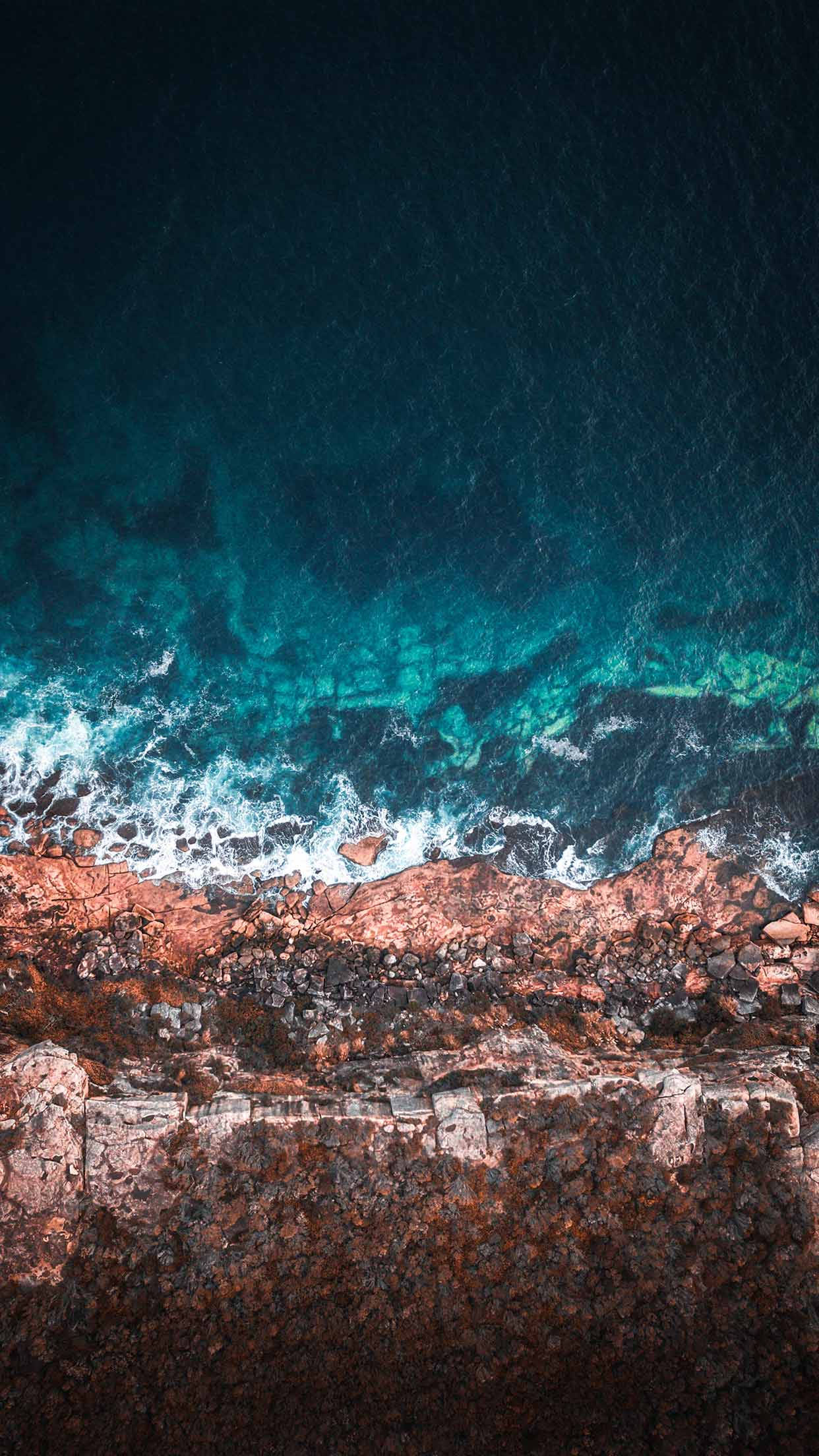 iPhone XS 深藍色海洋壁紙 壁紙 — 深藍色天空下，碧綠色的海浪拍打著崎嶇的紅褐色海岸懸崖的航拍視圖。