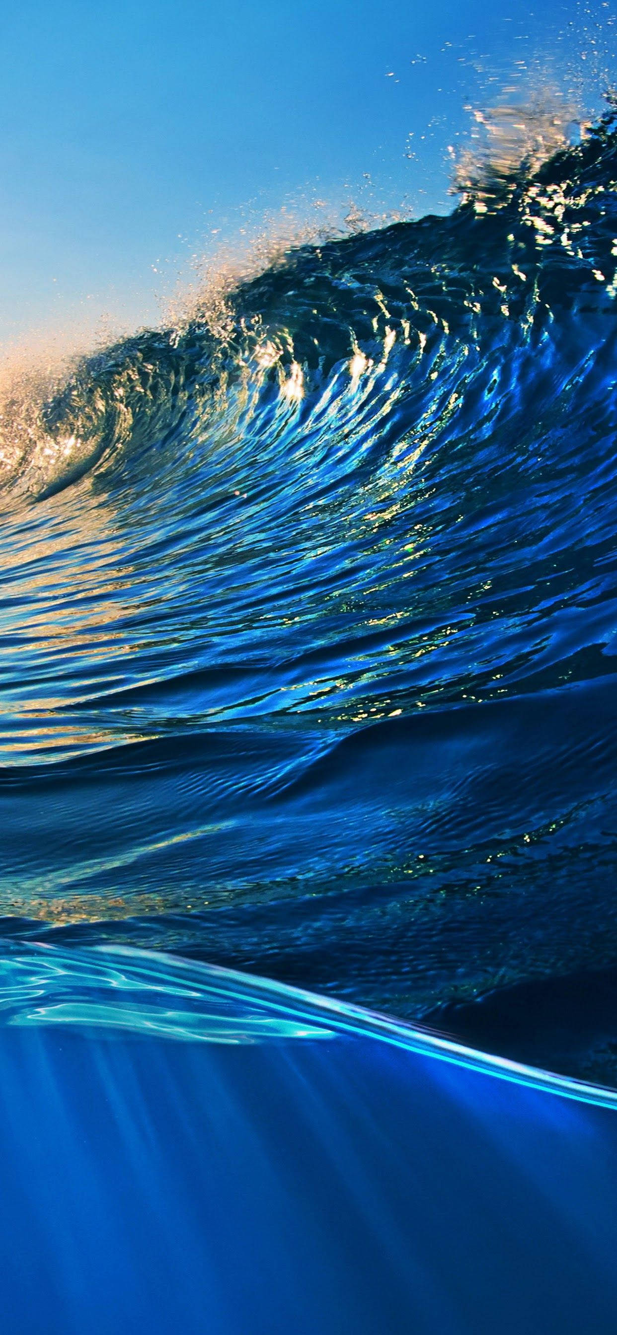 iPhone XS 深藍海浪 壁紙 — 一道高聳的海浪在晴朗的藍天下翻捲，陽光照射的水花四濺，下方是深藍色的海水。