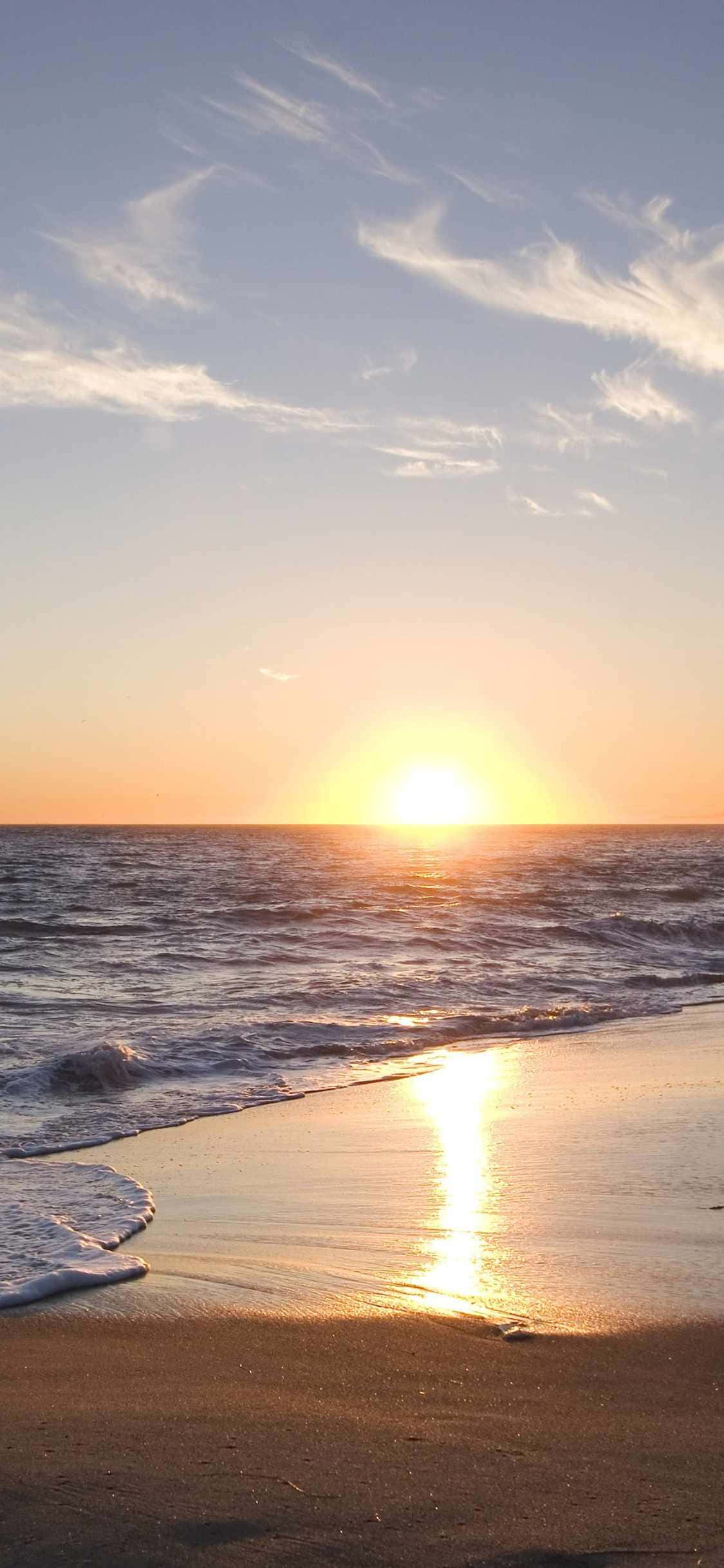 沉浸於加州馬里布美景的iPhone Xs影像 壁紙 — 金色夕陽照耀在海浪上，海浪沖刷著薄雲下的沙灘。