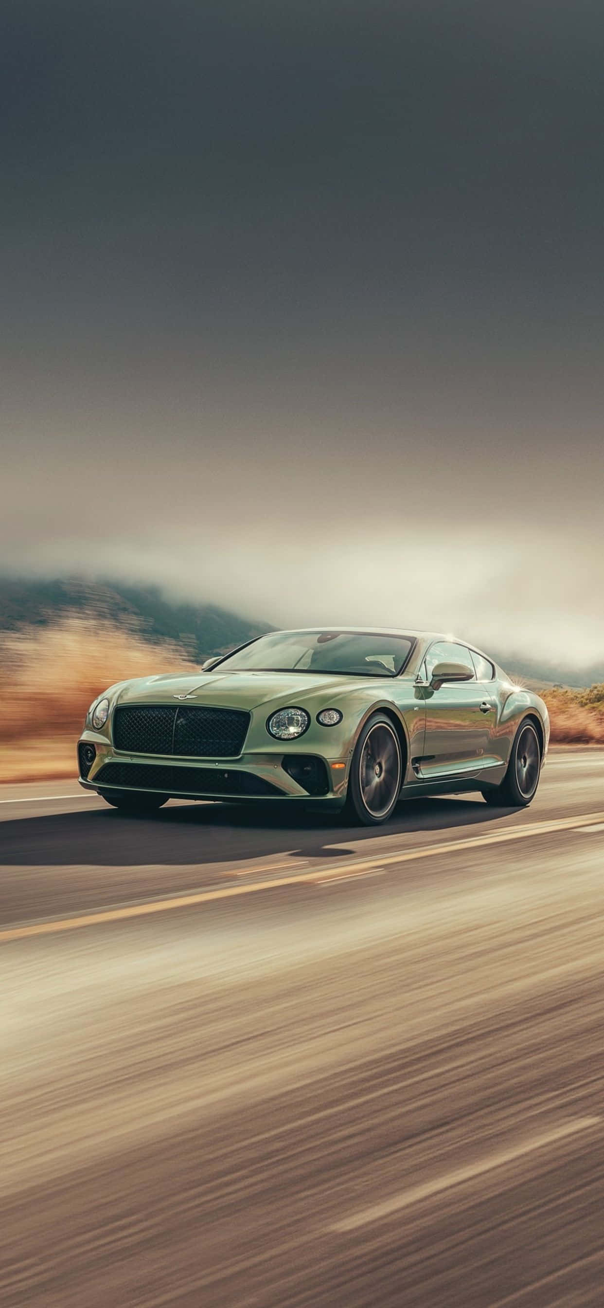 獨特高級：iPhone XS Max Bentley 壁紙 — 一輛時尚的綠色 Bentley Continental GT 在沙漠公路上疾馳，背景是模糊的動態和朦朧的山巒。