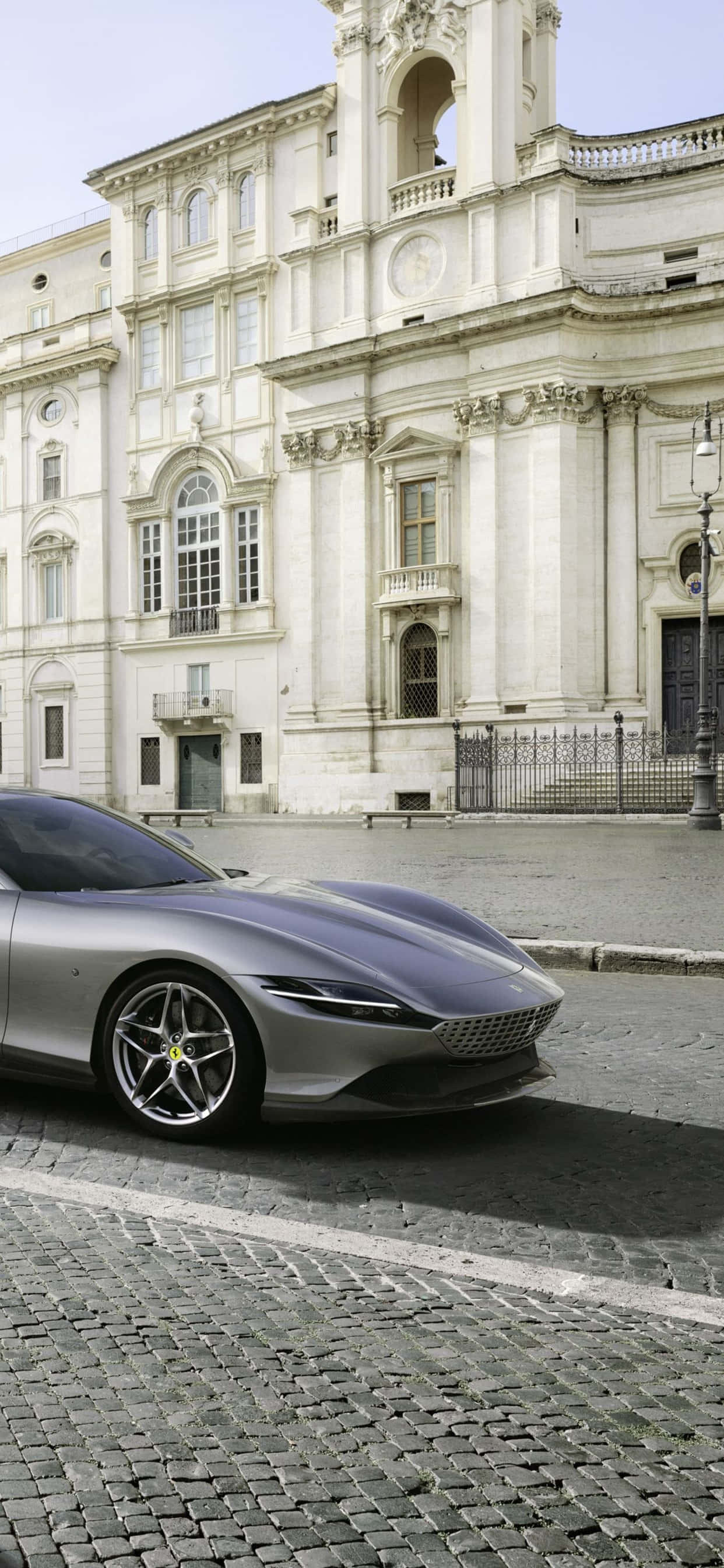 iPhone XS Max 賽車主題灰色 Ferrari Roma 背景 壁紙 — 一架時尚的銀色跑車停泊在鵝卵石街道上，前面是一座宏偉的古典建築。