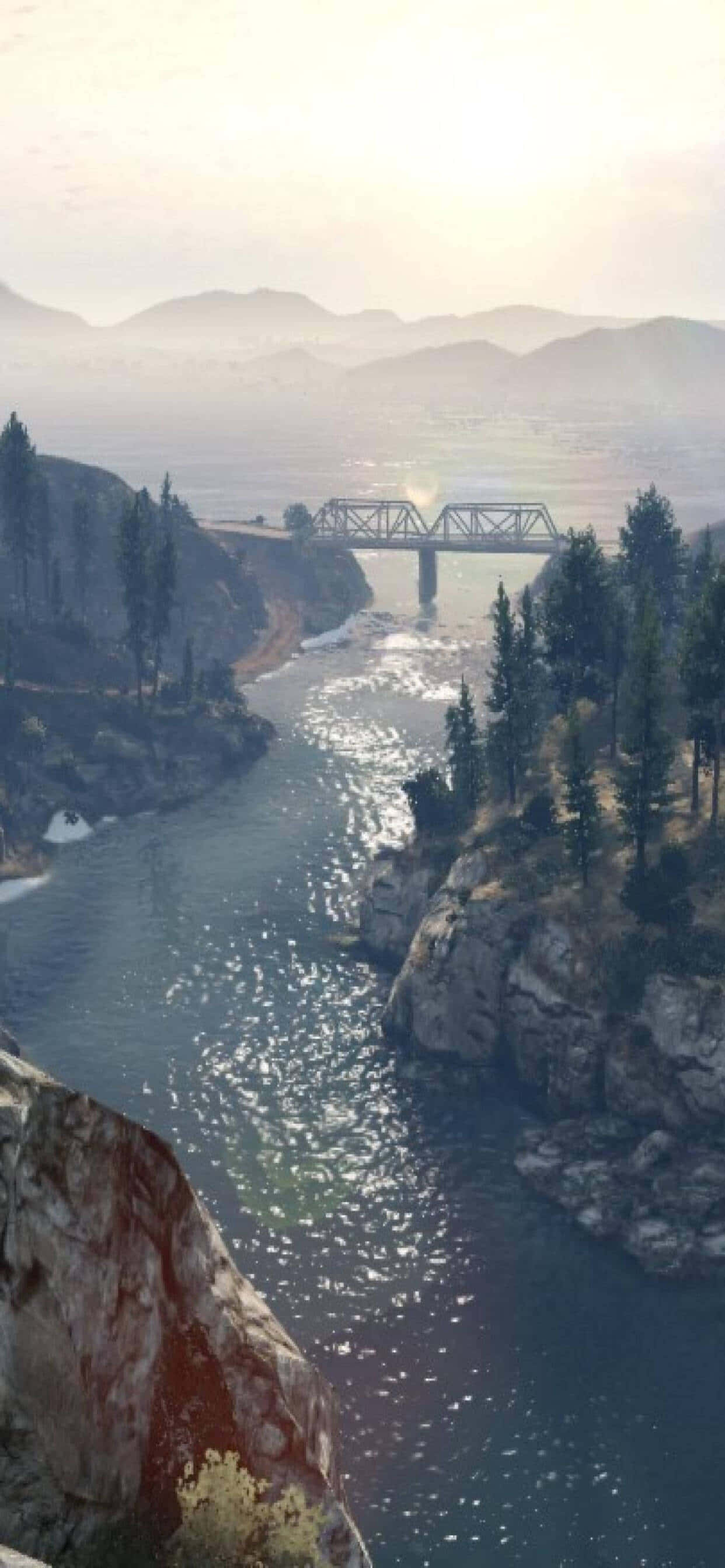 iPhone XS Max《GTA V》洛聖都河景背景 壁紙 — 一座鋼桁架橋橫跨蜿蜒的河流，穿過霧氣瀰漫的山巒，松林和岩石峭壁在柔和的金色光芒下。