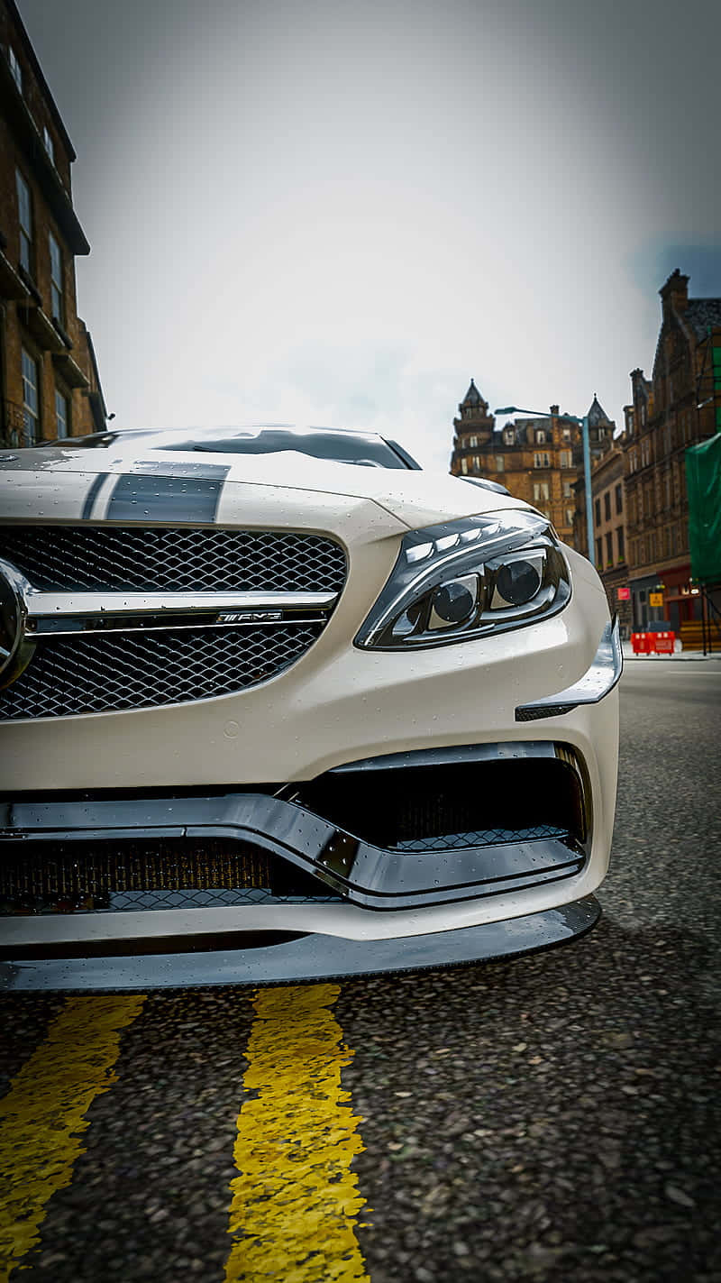 iPhone XS Max Project Cars 2 白色梅賽德斯-奔馳 E 背景 壁紙 — 一輛白色 Mercedes-AMG 跑車停泊在濕滑的城市街道上，背景有黃色道路標線和歷史建築的低角度拍攝。