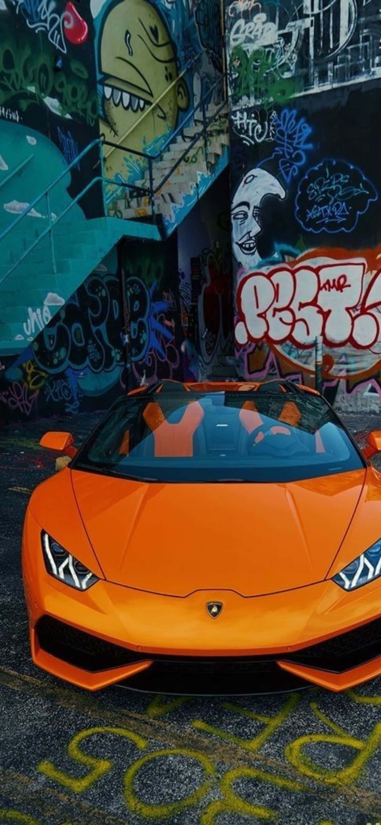 iPhone XS Max 橙色蘭博基尼Huracan 背景 壁紙 — 一輛充滿活力的橙色林寶堅尼跑車停泊在佈滿塗鴉的都市小巷牆前。