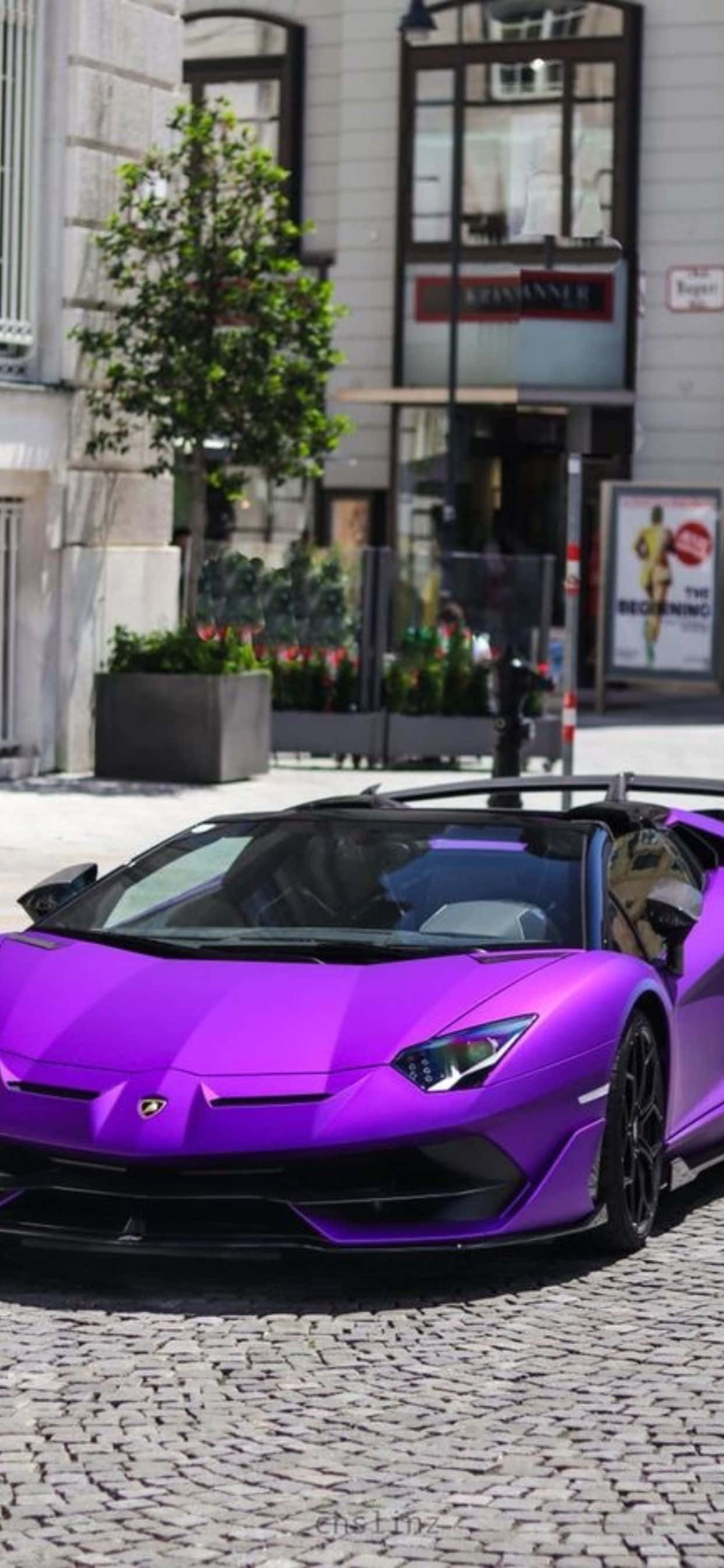 iPhone XS Max 紫黑蘭博基尼Aventador 背景 壁紙 — 一架充滿活力的紫色林寶堅尼 Aventador 開篷跑車停泊在鵝卵石街道上，前方是一座帶有綠植的現代建築。