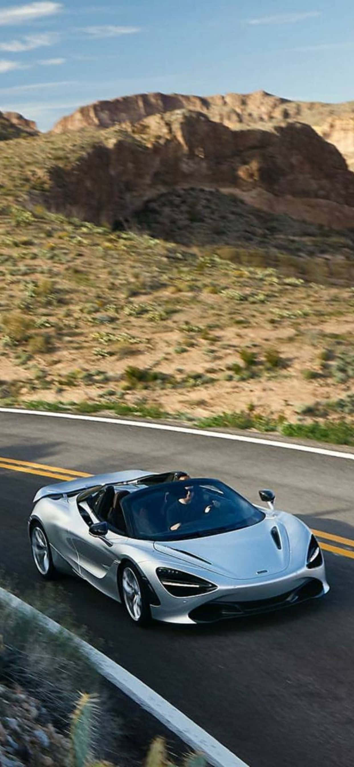 iPhone XS Max 與 McLaren 720S 背景 — Supernova 銀色 壁紙 — 一輛銀色McLaren跑車在晴朗藍天下沿著蜿蜒的沙漠道路行駛，兩旁是崎嶇的紅色岩層。