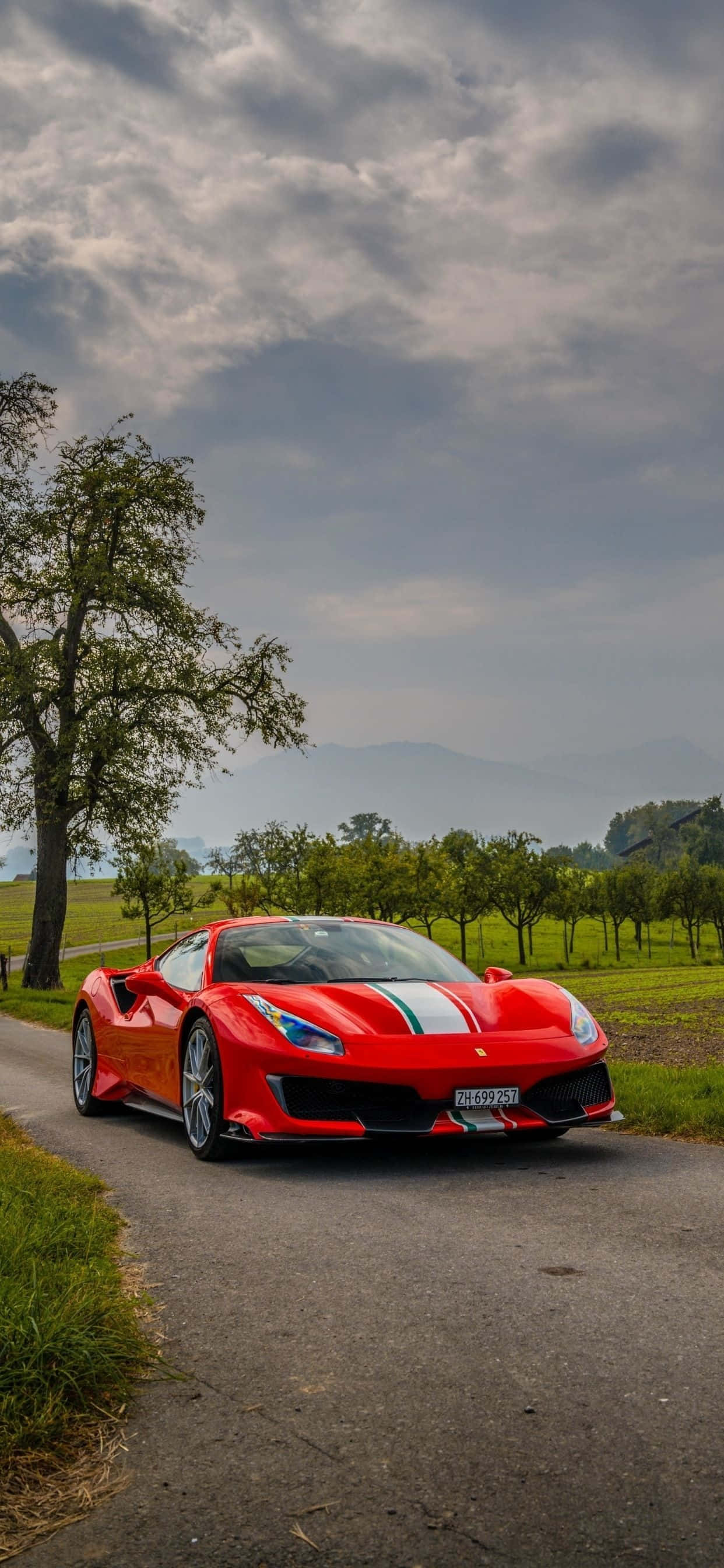 iPhone XS Max 賽車背景：紅色法拉利 488 Pista 壁紙 — 一輛帶有白色條紋的鮮紅色法拉利跑車，停泊在蜿蜒的鄉村道路上，天空多雲。