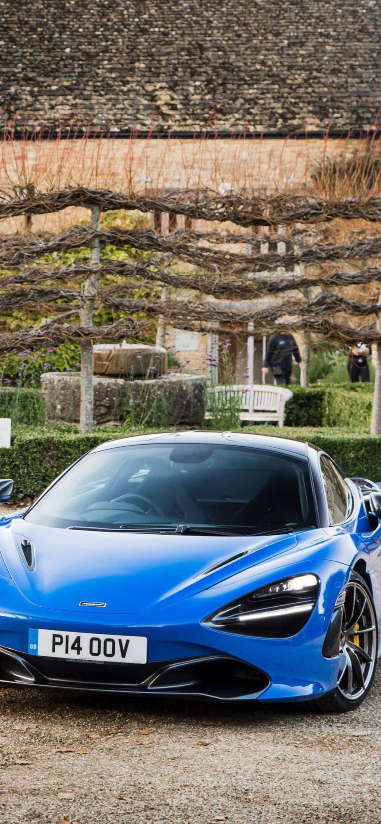 iPhone XS Max 靜置於 McLaren 720S 之上 壁紙 — 一輛充滿活力的藍色McLaren跑車停在碎石路上，下方是鄉村風格的葡萄園涼棚，周圍有石牆和修剪整齊的樹籬。