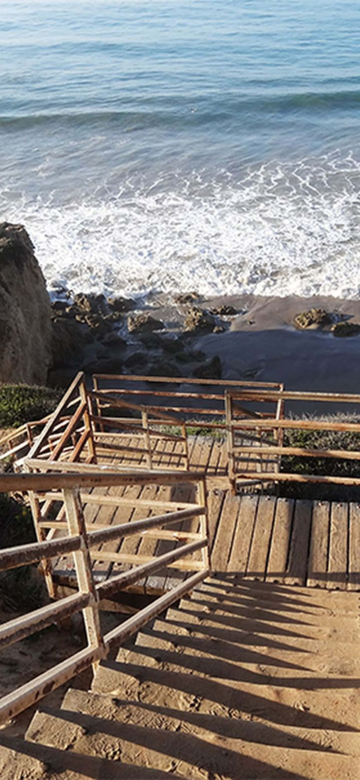 iPhone Xs Max 馬利布海灘階梯背景 壁紙 — 木樓梯向下通往岩石海灘，海浪在明亮的陽光下拍打。