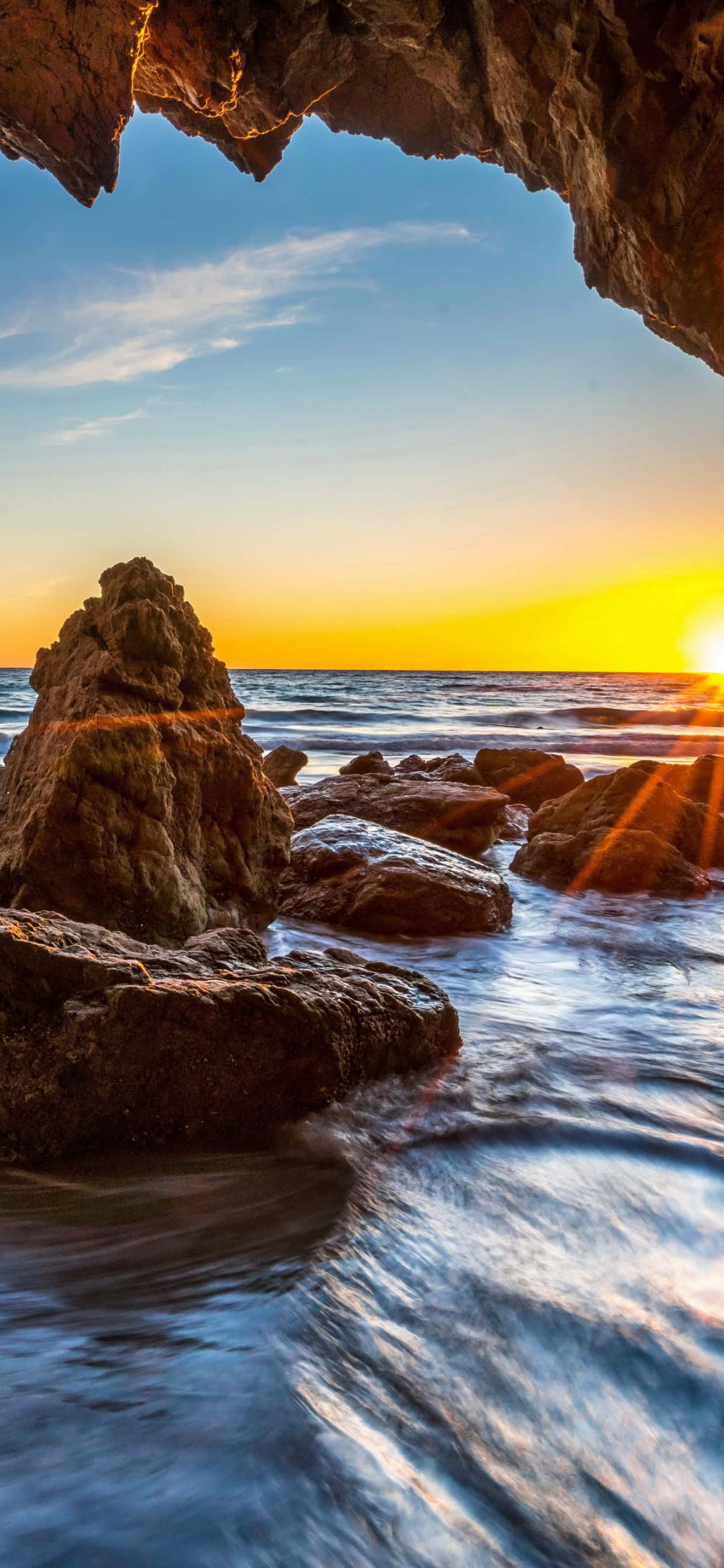 iPhone Xs Max 馬利布海灘洞穴旁岩石背景 壁紙 — 夕陽穿過岩石洞穴開口發光，海浪在海洋邊緣的巨石上流過。