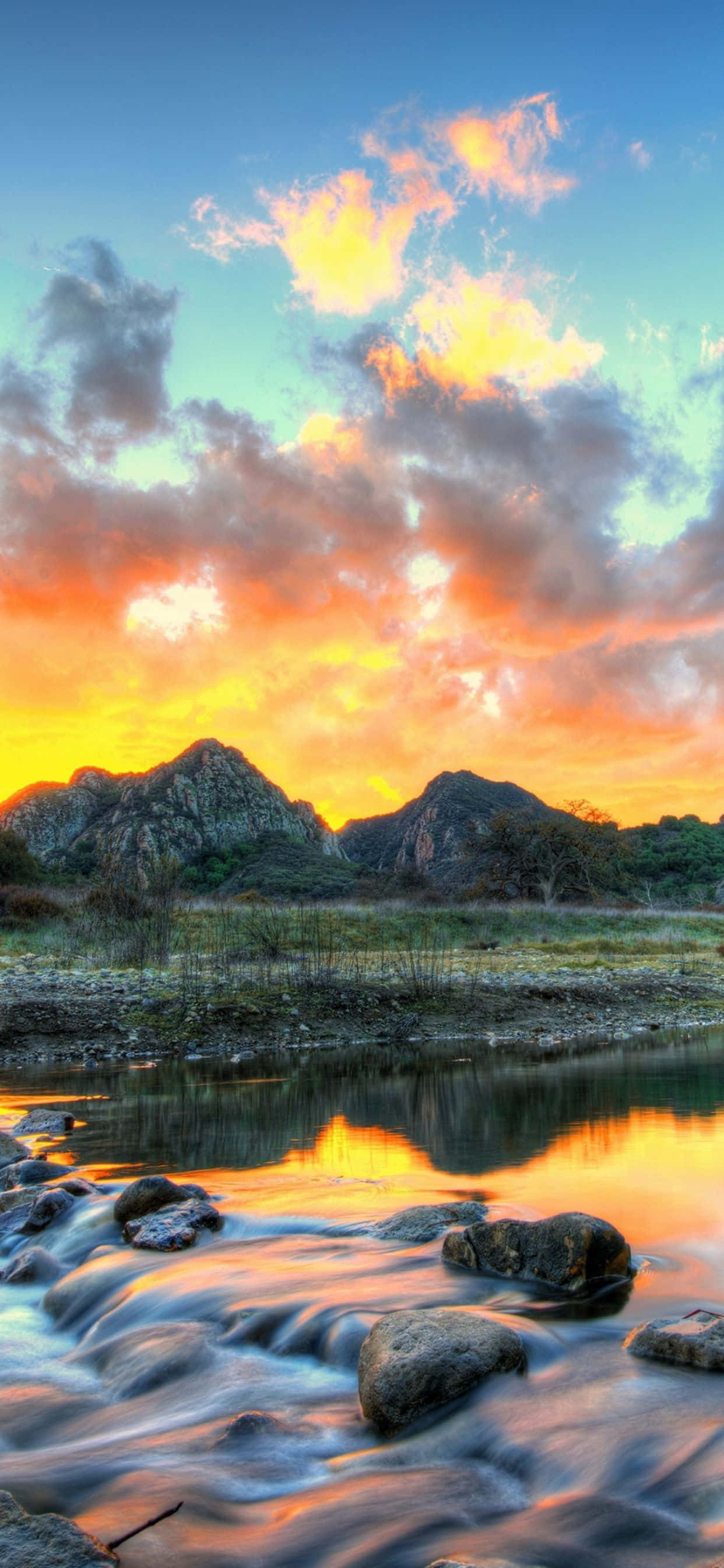 iPhone XS Max 馬里布倒影河流背景 壁紙 — 鮮豔的夕陽以火熱的橙色和粉紅色調繪製天空，映照在崎嶇的山脈上，並反射在一條流動的河流中，河水平滑，岩石散落。