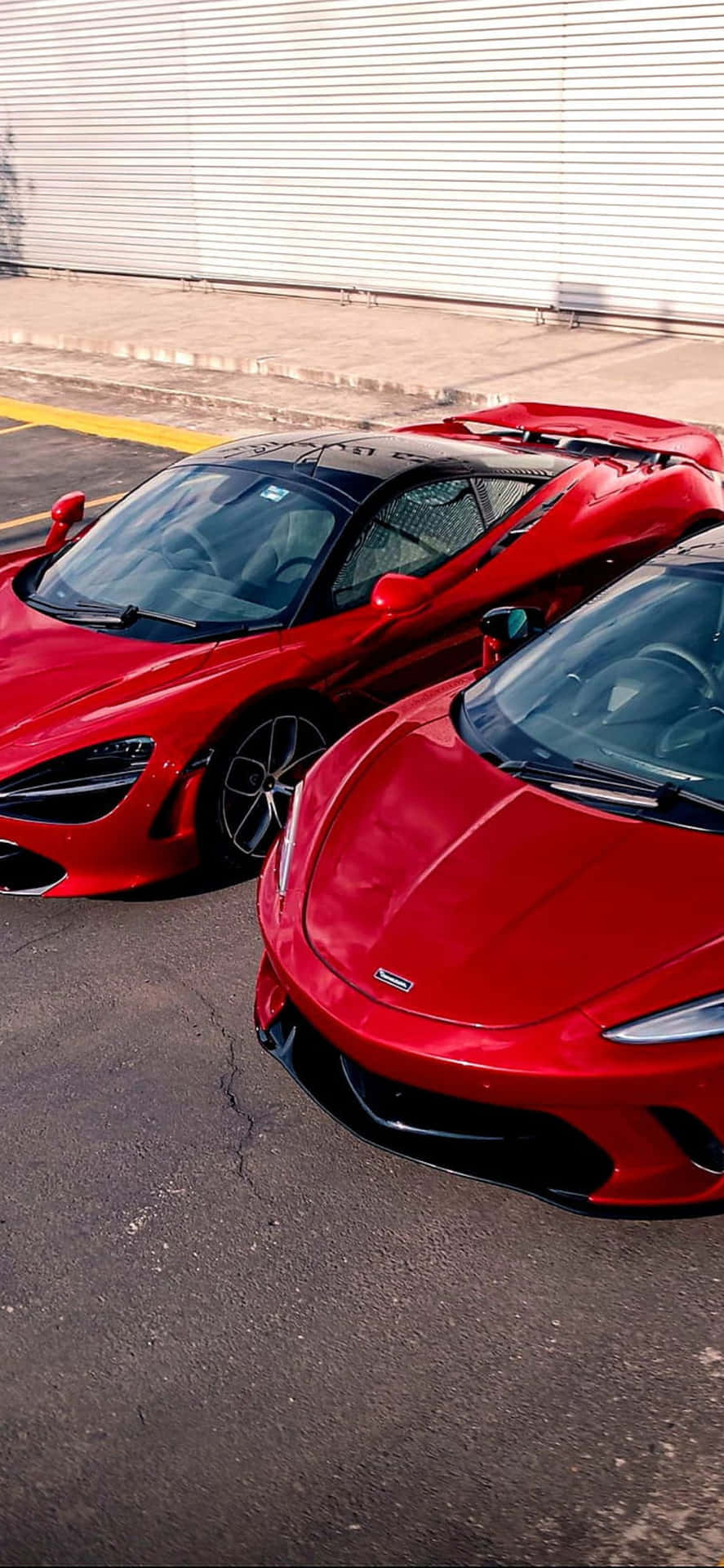 iPhone Xs Max與McLaren 720S - 梅賽德斯紅色背景 壁紙 — 兩輛亮面紅色McLaren超級跑車並排停泊在瀝青路面上，旁邊是波紋金屬牆。