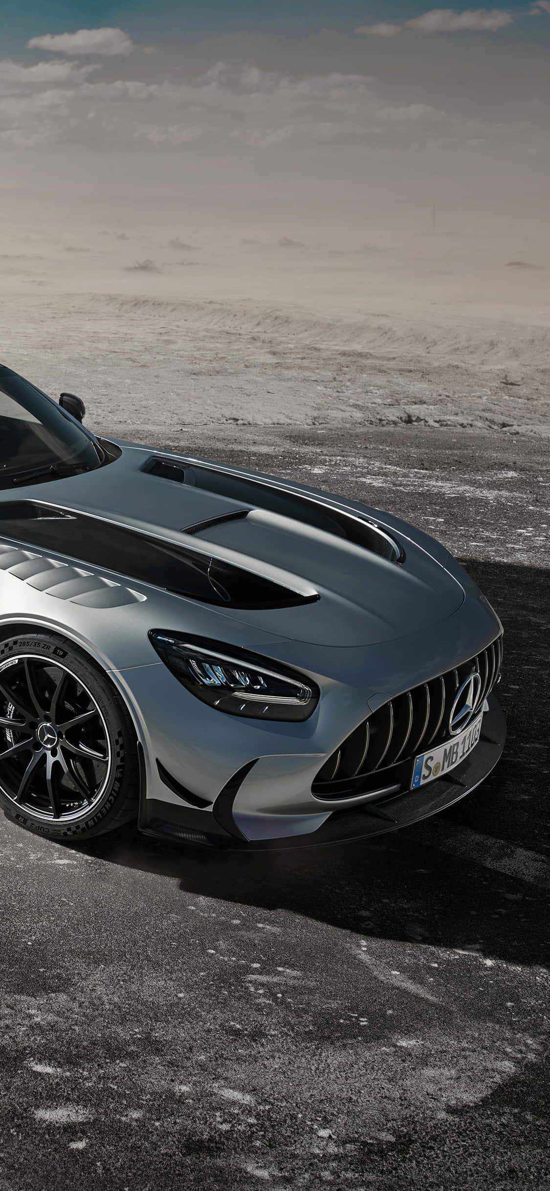 銀色 iPhone XS Mercedes AMG 沙漠邊緣背景 壁紙 — 一架流線型銀色 Mercedes-AMG GT R 跑車停泊在積雪瀝青路上，天空多雲。