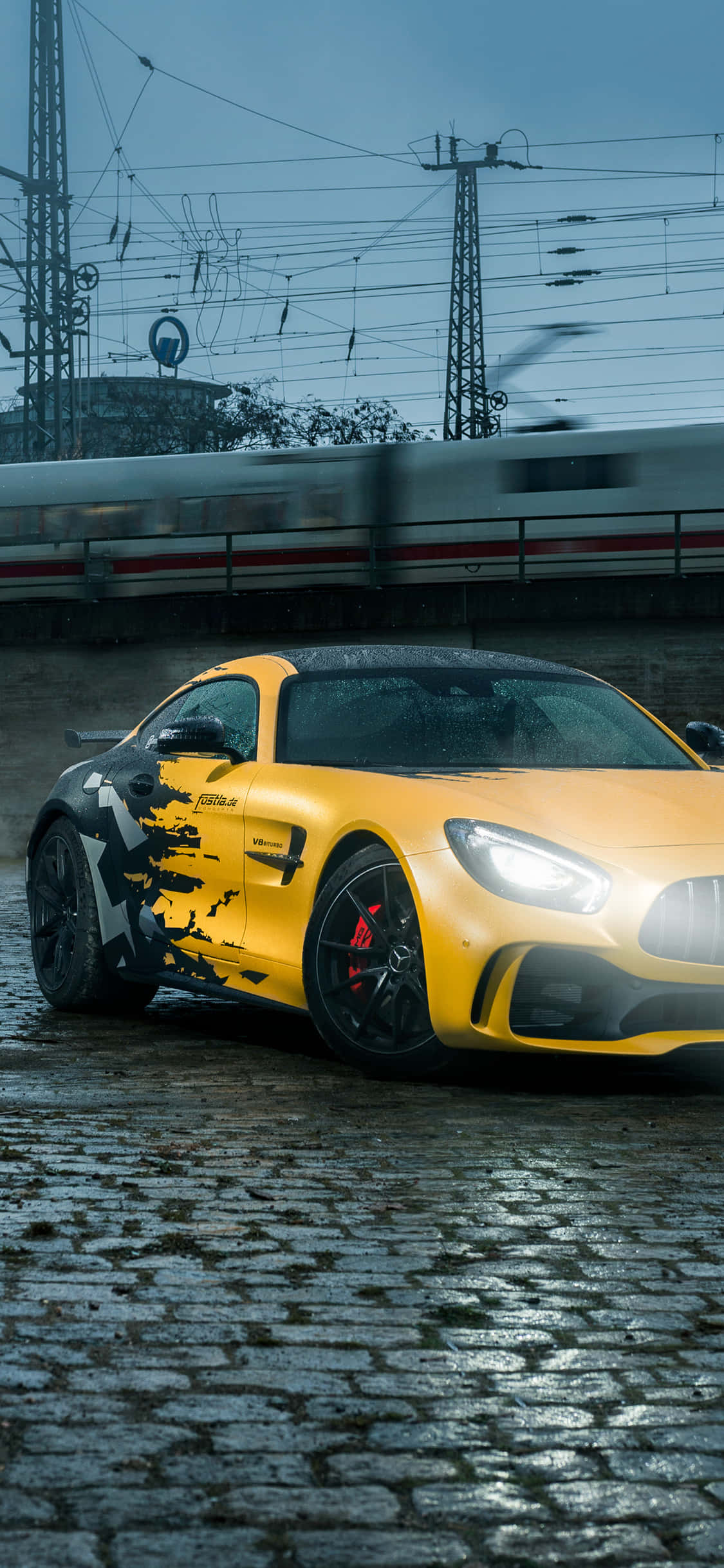 黑色與黃色 iPhone XS Mercedes AMG 背景 壁紙 — 一輛帶有黑色潑濺圖案的鮮黃色跑車停在濕漉漉的鵝卵石上，一列模糊的火車在陰沉的暮色天空下高速駛過。