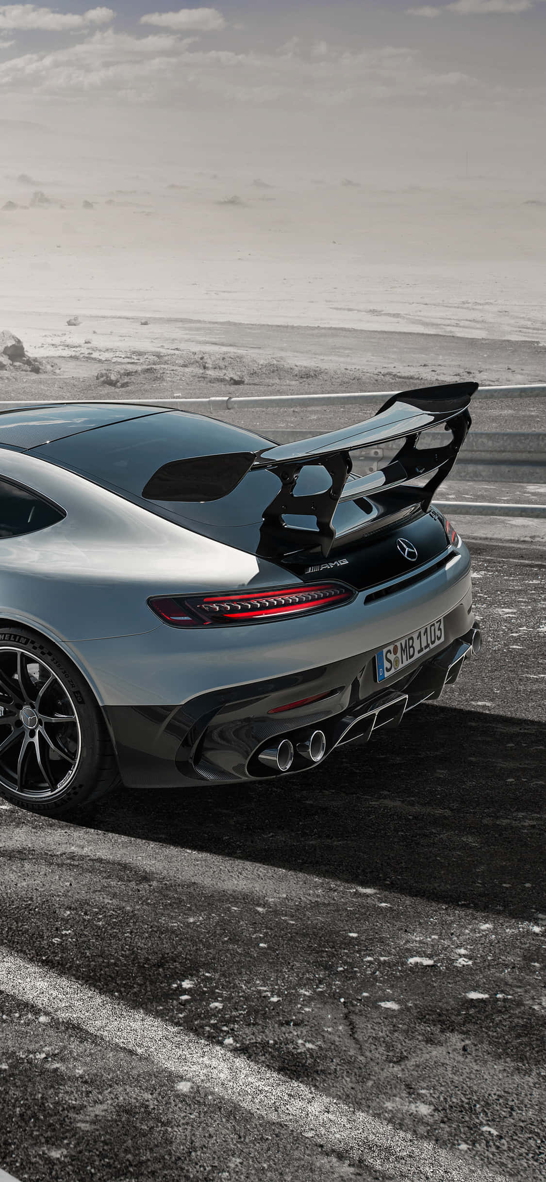 iPhone XS 捕捉的 Mercedes AMG 崇高風景背景 壁紙 — 一架銀色梅賽德斯-AMG GT R跑車配備大型尾翼，停泊在積雪海岸線附近潮濕的瀝青路上，天空多雲。