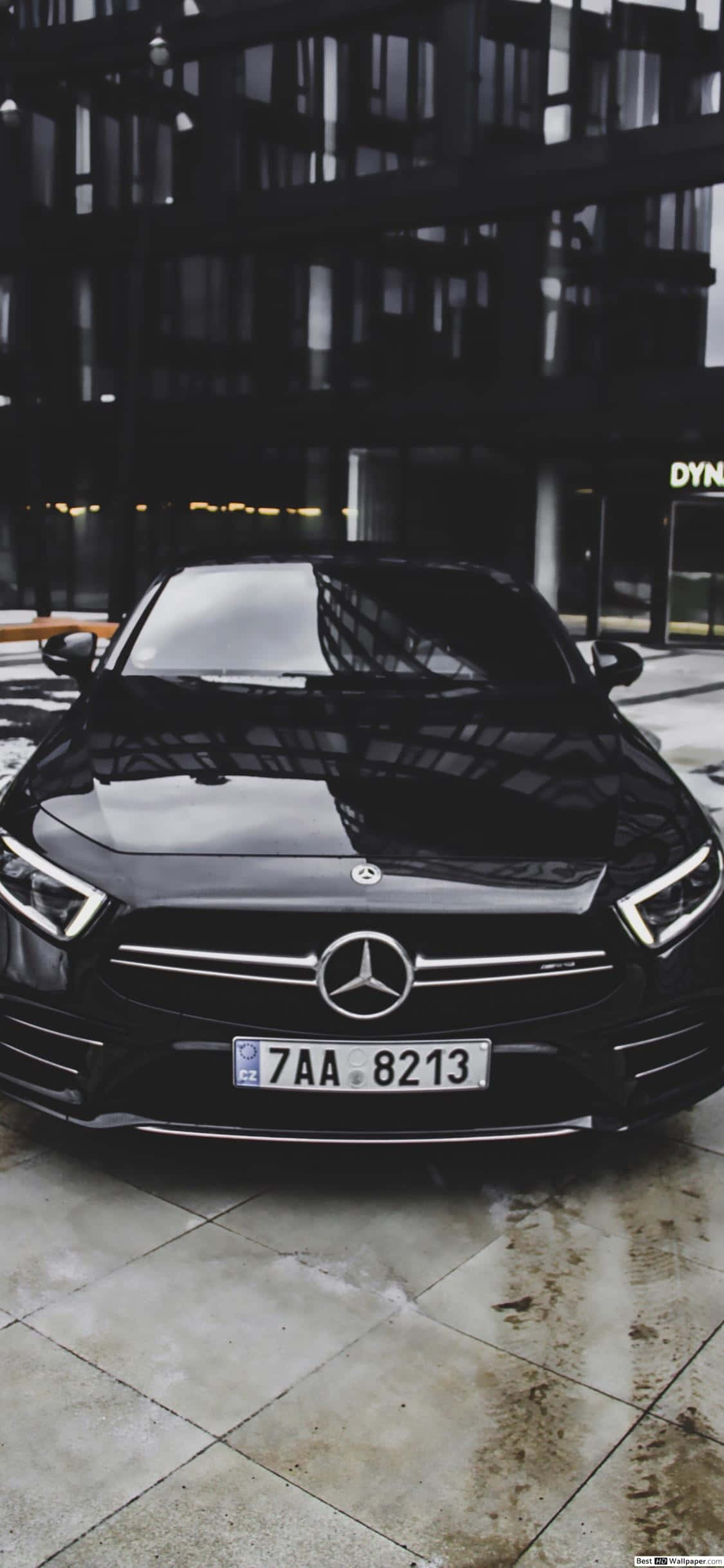 iPhone XS 背景：黑色 Mercedes-Benz CLS 壁紙 — 一輛時尚的黑色平治房車在夜間停泊在現代玻璃建築下的濕滑路面上。