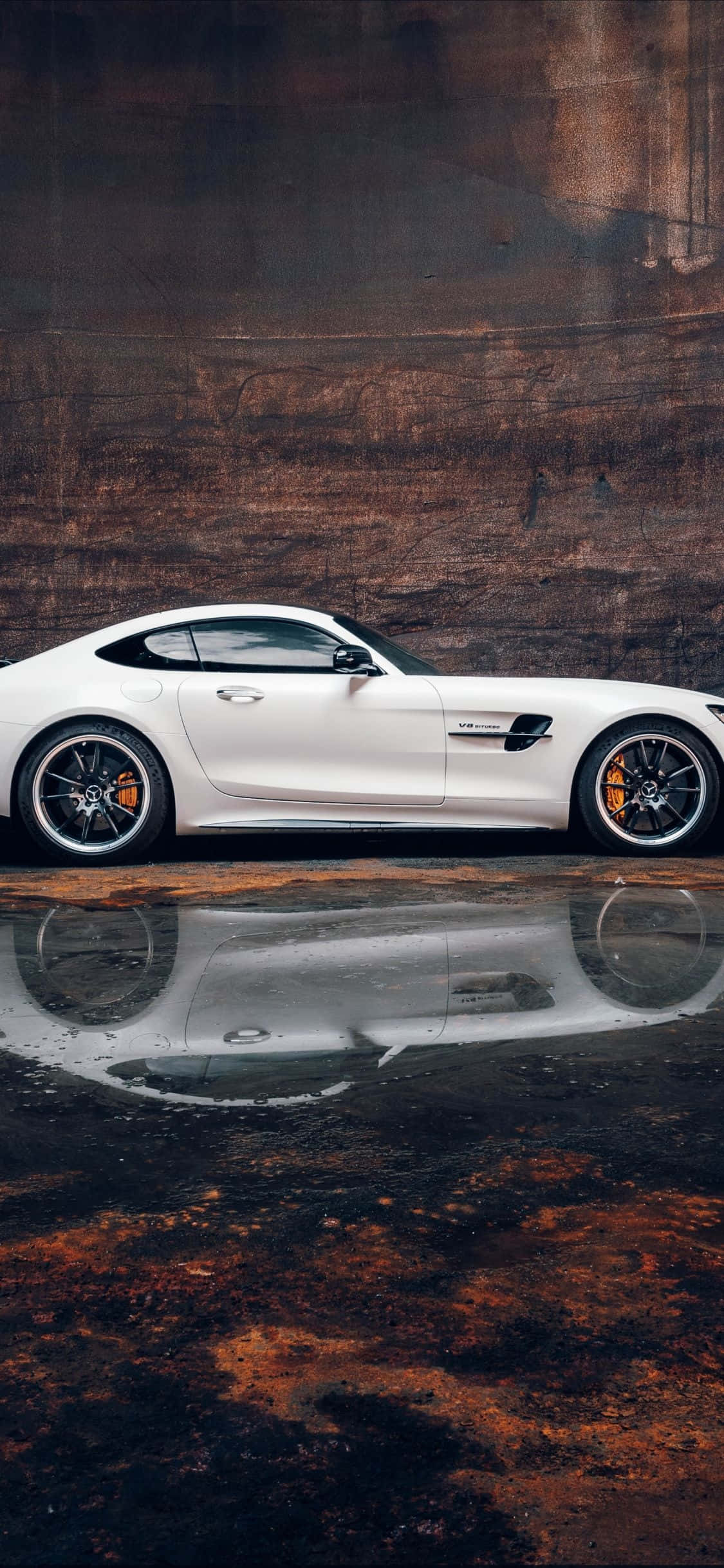 iPhone XS 背景：白色梅賽德斯-奔馳 SLS AMG 壁紙 — 一輛時尚的白色跑車在戲劇性燈光下，映照在濕漉漉的路面上，背景是質感粗糙的鏽蝕牆壁。