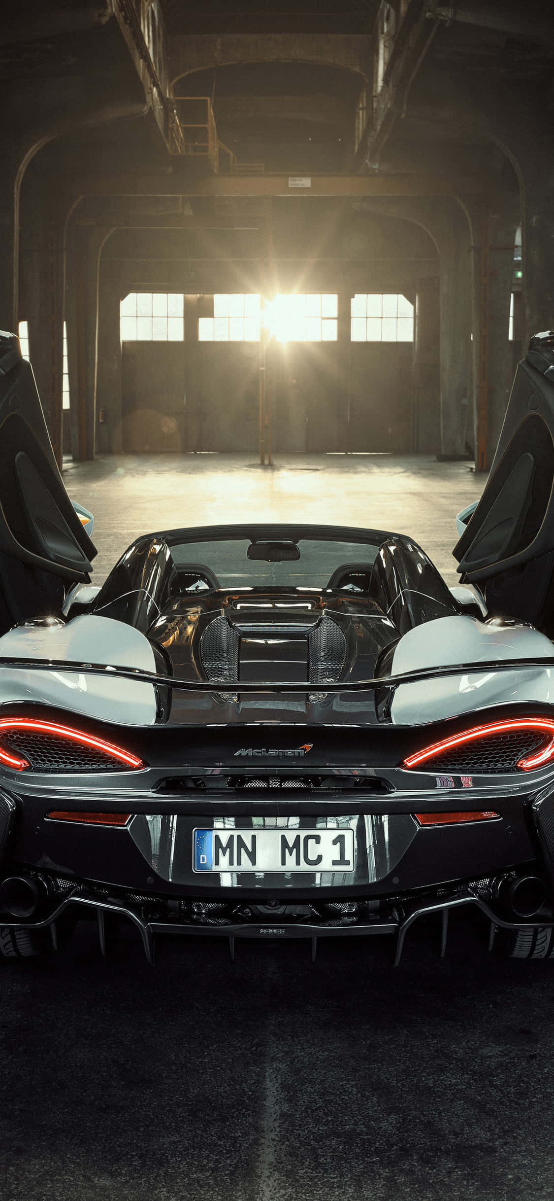iPhone Xs 在 McLaren 720S 前顯得格外合拍 壁紙 — 一架時尚的黑色McLaren跑車，帶有發光的紅色尾燈，停放在陽光普照的工業倉庫中，鷗翼車門敞開。