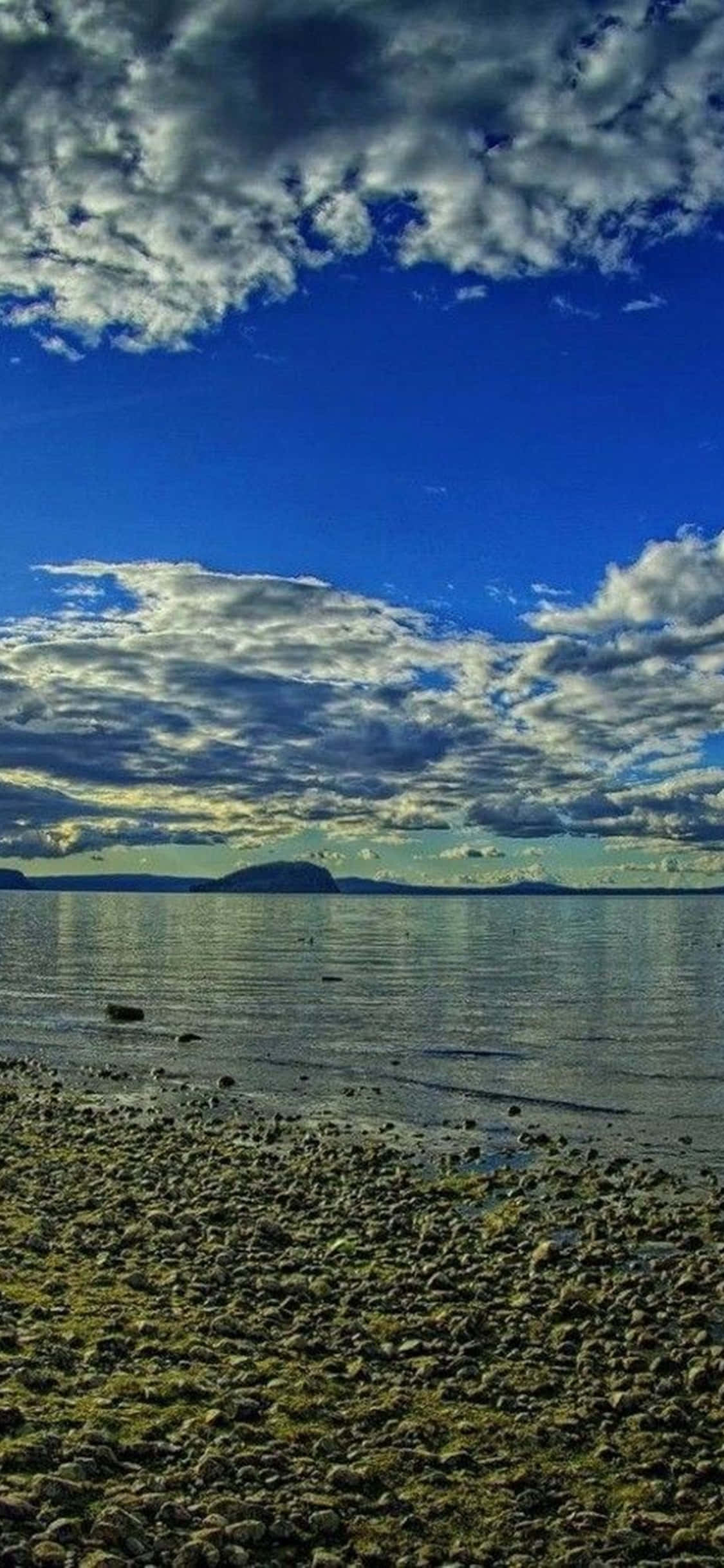 iPhone XS 桌面 PC 海灘石子背景 壁紙 — 一片充滿活力的藍天，帶有戲劇性的雲彩，倒映在平靜的水面上，與岩石海岸相接。