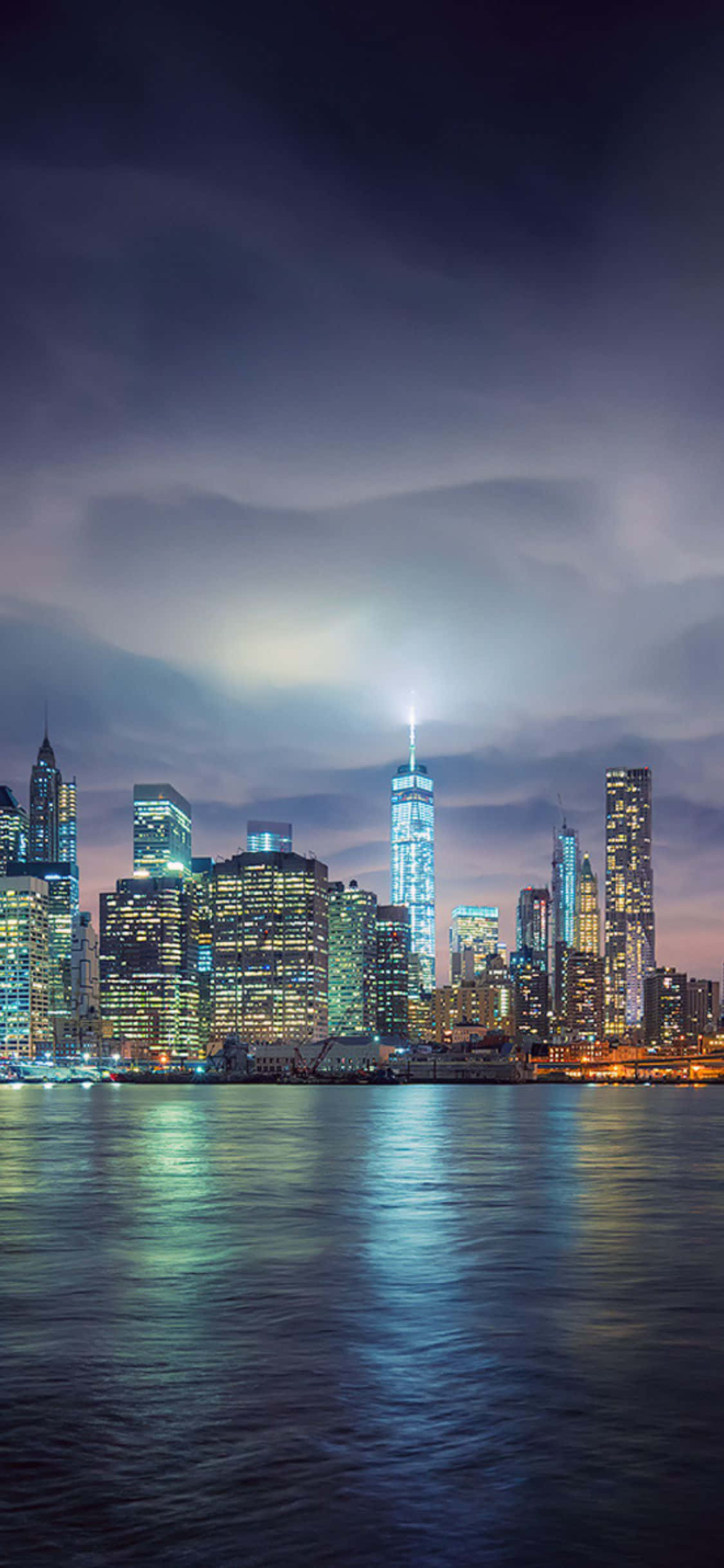iPhone Xs 眺望紐約市天際線 壁紙 — 充滿活力的紐約天際線夜景，倒映在平靜的水面上，籠罩在憂鬱的暮色天空下。