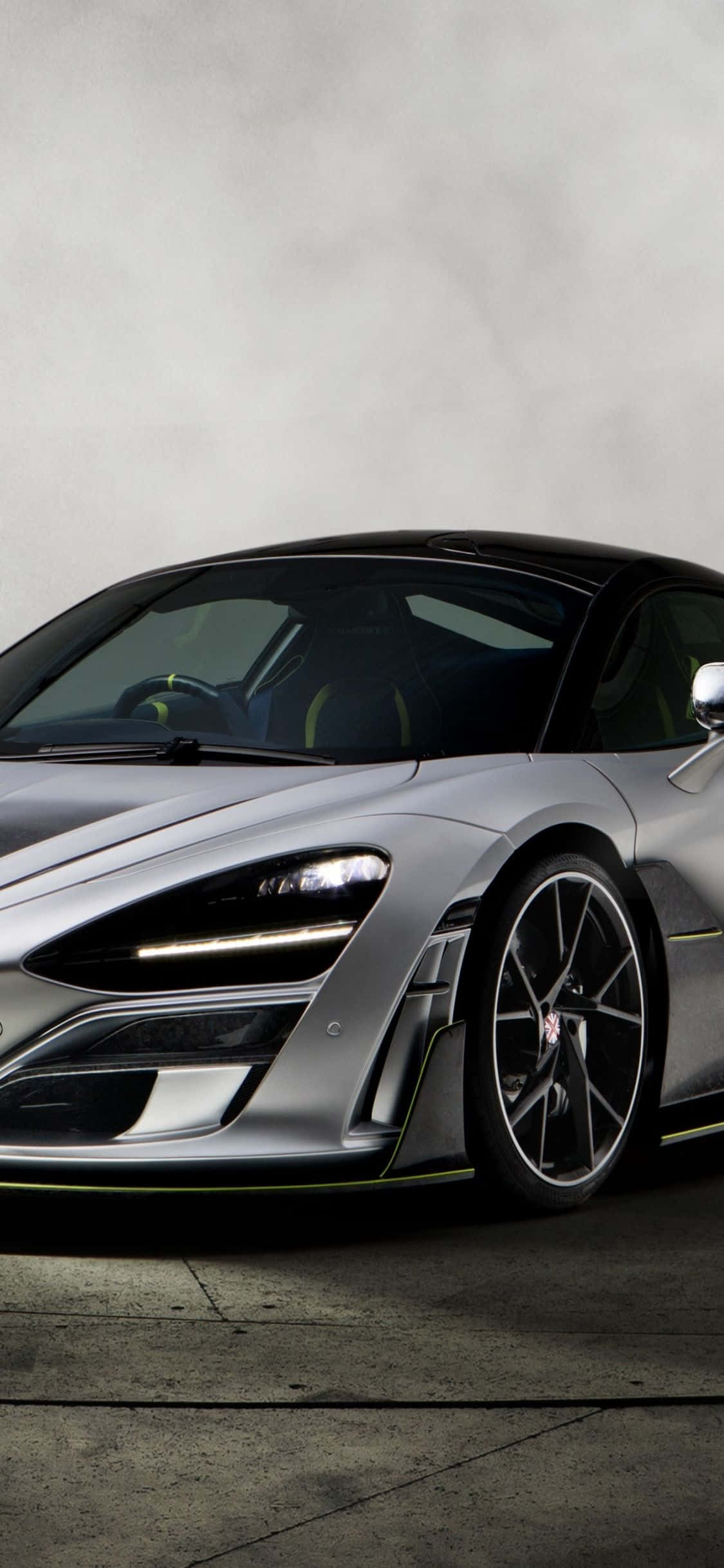 iPhone Xs 與 McLaren 720S 的奢華聯名 壁紙 — 一輛時尚的銀色跑車，帶有發光頭燈和黑色飾邊，停在混凝土表面上，背景是柔和的灰色牆壁。