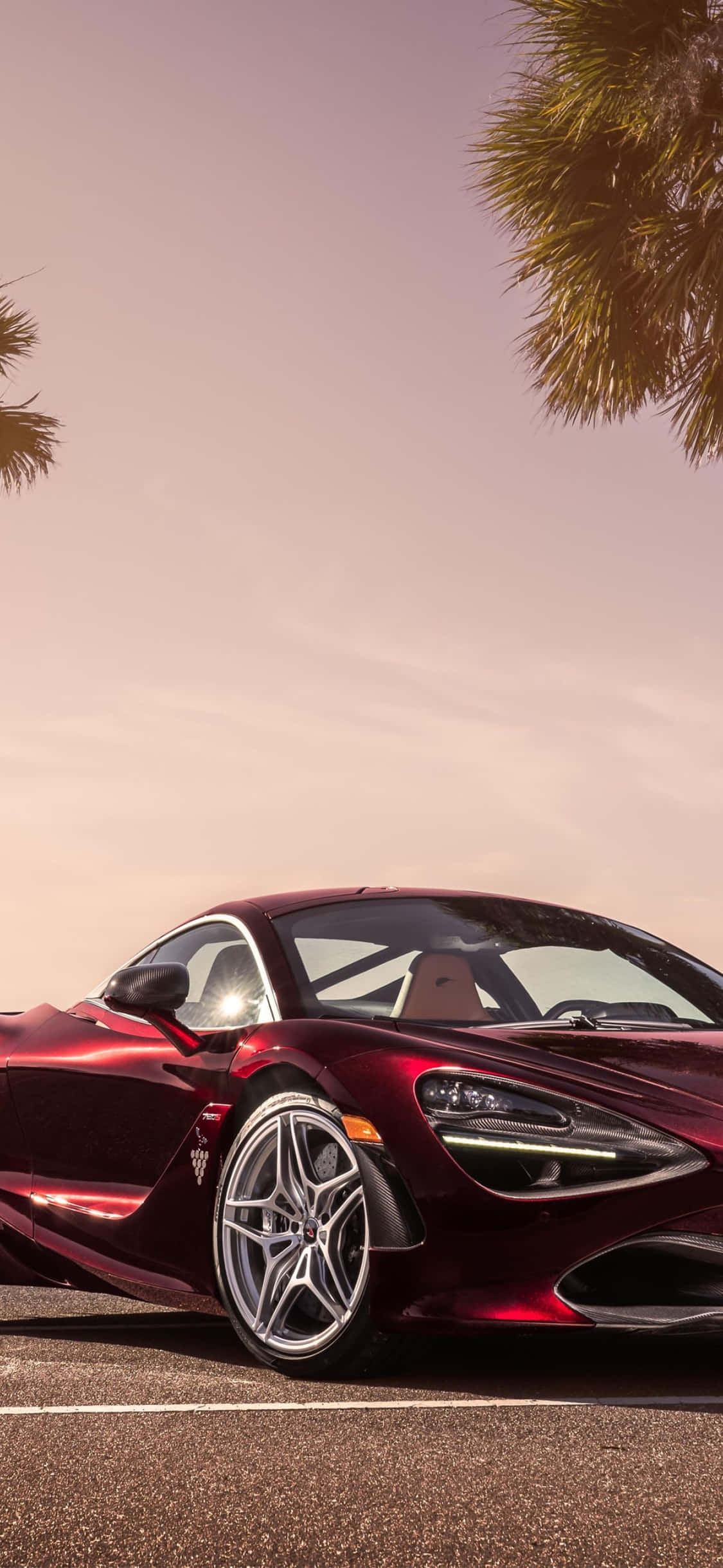 iPhone Xs 與 McLaren 720S：奢華的象徵 壁紙 — 一輛時尚的酒紅色跑車停在棕櫚樹下，夕陽西下，天空溫暖閃耀。