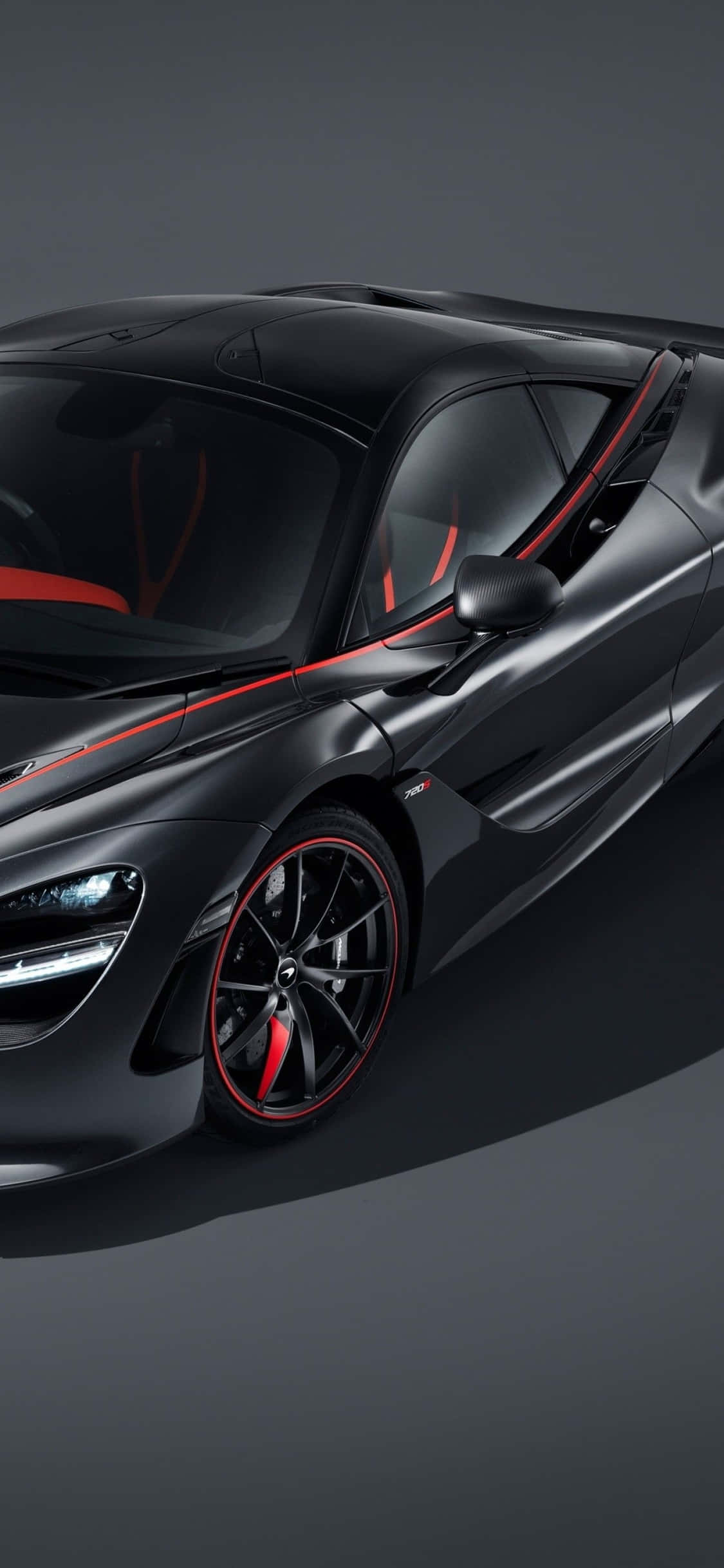 iPhone Xs 與流線型 McLaren 720s 的完美搭配 壁紙 — 一輛時尚的啞光黑色McLaren跑車，帶有紅色點綴和發光的車頭燈，以深灰色工作室背景為襯托。