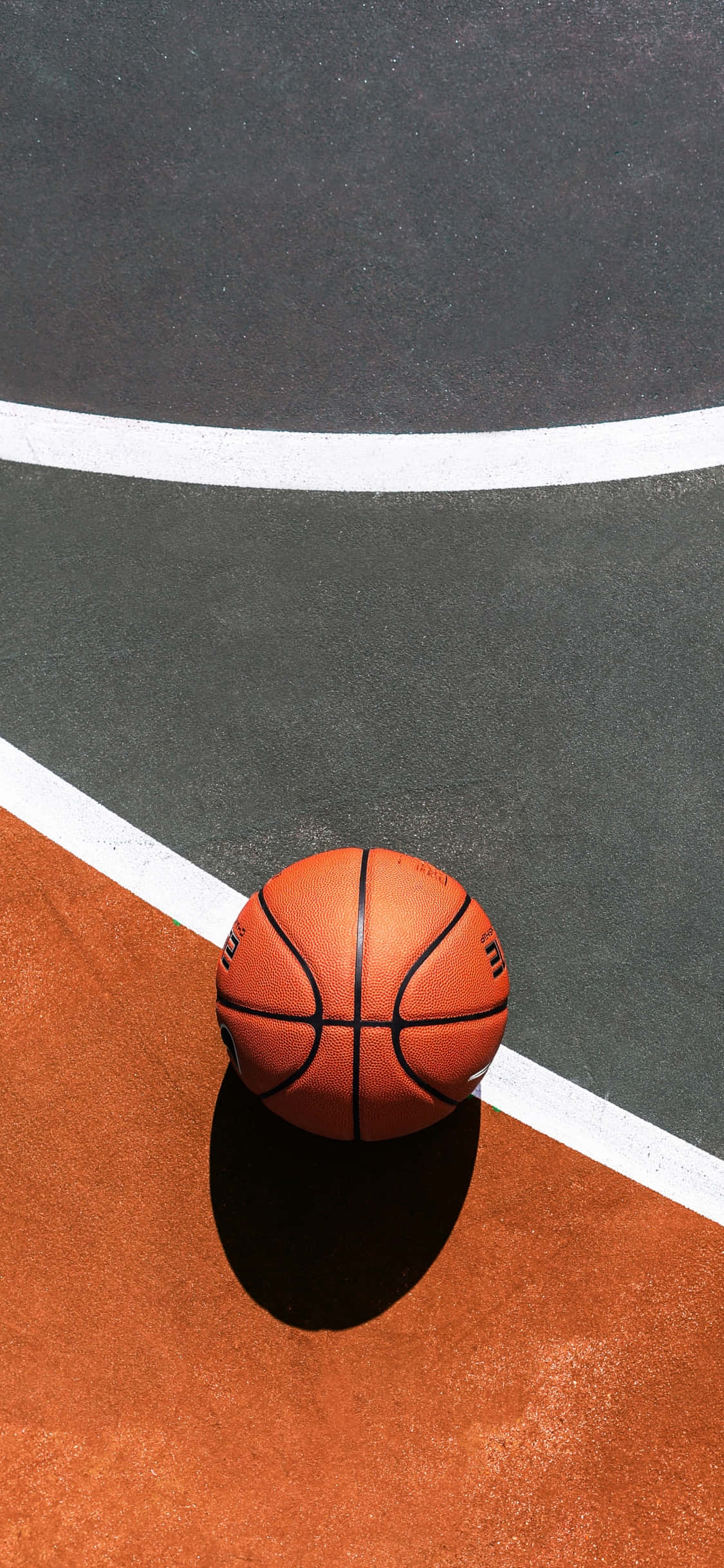 iPhone Xs籃球NBA選秀背景 壁紙 — 一個籃球放在陽光普照的戶外球場上，球場有清晰的白色線條和帶紋理的橙色及灰色表面。