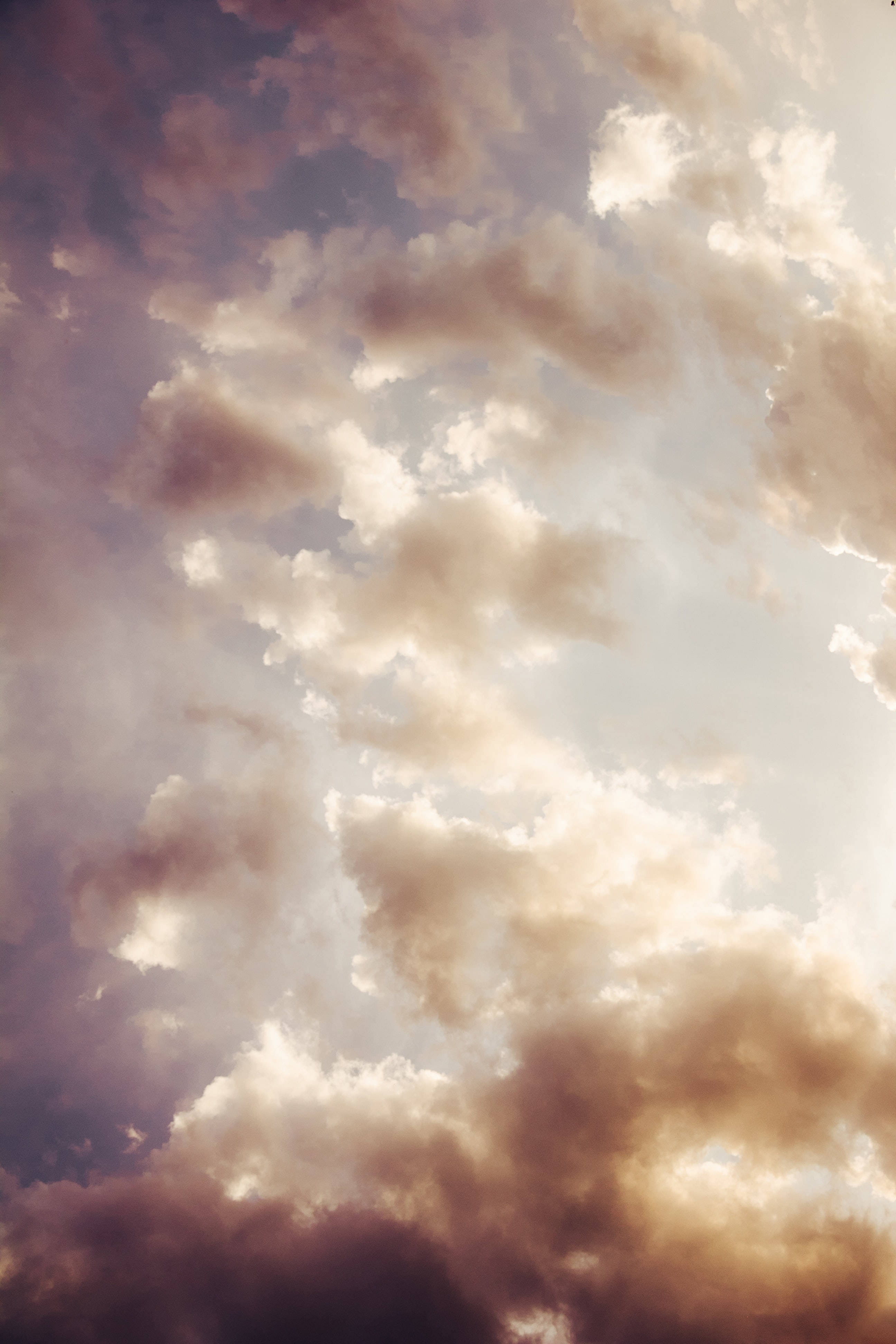 天際雲 iPhone 壁紙 — 戲劇性的雲朵以溫暖的深褐色調在柔和的天空中飄移，帶有金色亮點和深紫色陰影。