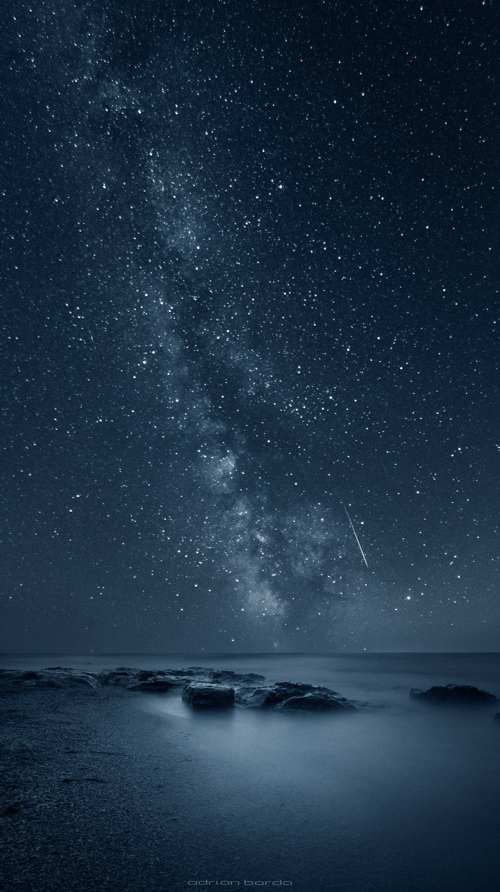 流星星際iPhone壁紙 壁紙 — 繁星點點的夜空，銀河與流星在平靜的海洋和前景的黑色岩石上拱起。