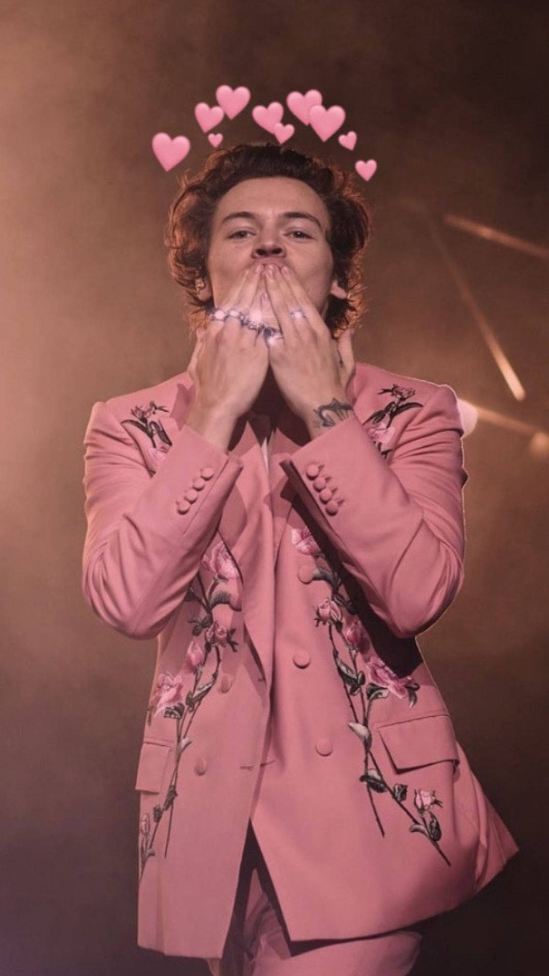 Harry Styles 身穿粉紅色花卉西裝，在舞台上飛吻，頭頂有漂浮的心形。