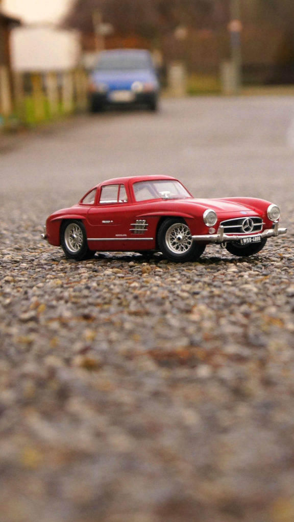 iPhone 安裝於汽車儀表板上 壁紙 — 一架鮮紅色復古Mercedes-Benz玩具車停泊在碎石路上，背景有一輛模糊的藍色汽車。