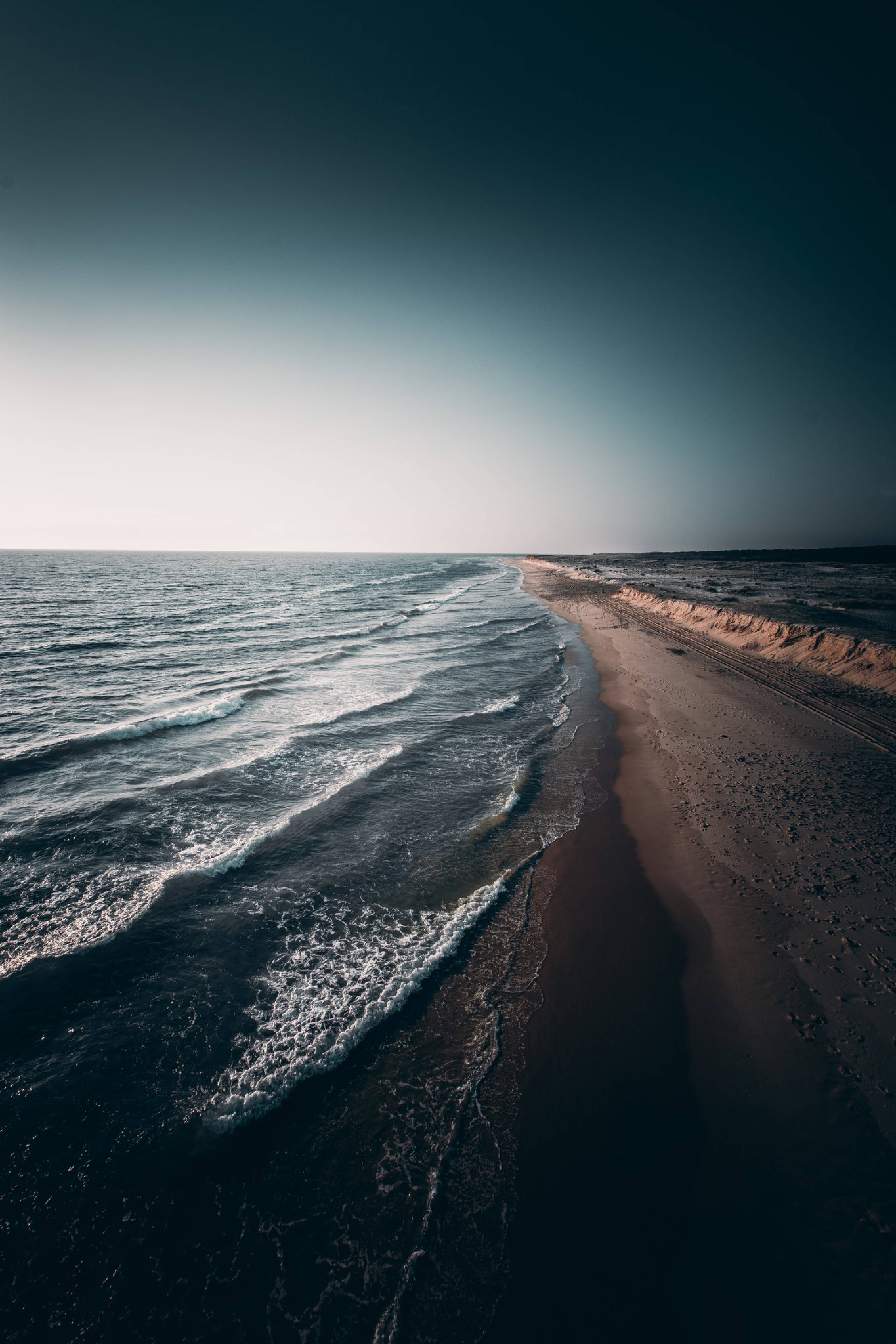 iPhone 海浪拍岸 壁紙 — 寧靜的海岸景色，在廣闊的暮色天空下，溫柔的海浪拍打著沙灘。