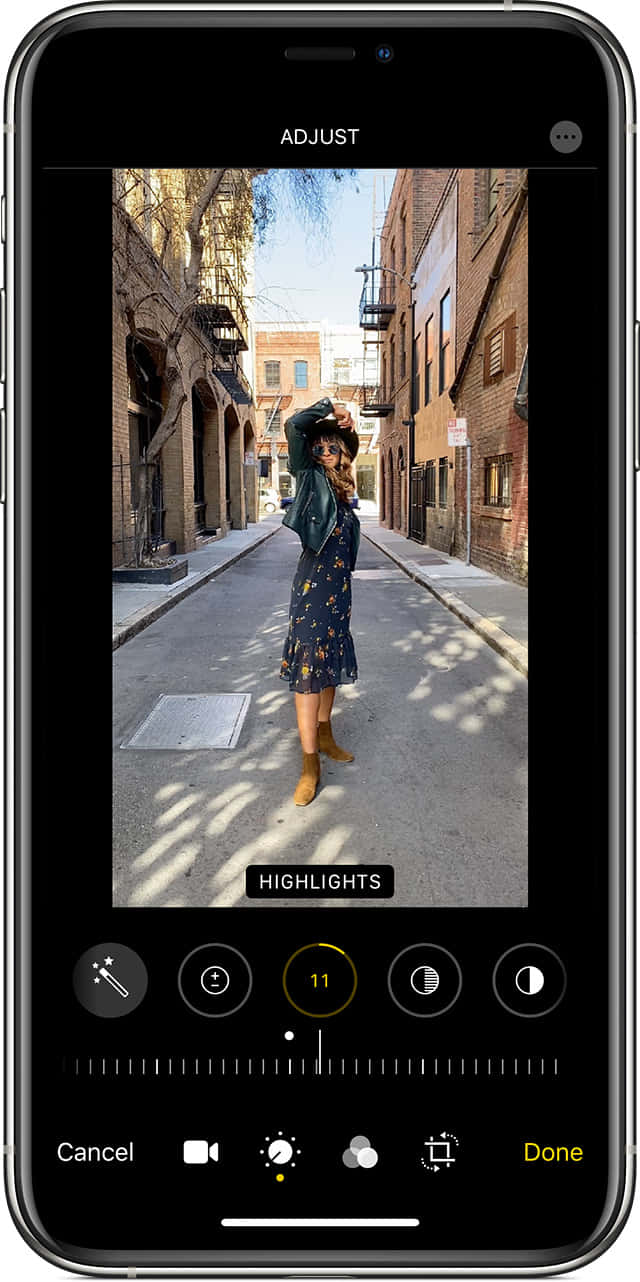 iPhone 的創新科技 壁紙 — 一位身穿花卉連衣裙和靴子的女性在磚砌建築之間的陽光普照的城市小巷中俏皮地擺姿勢。