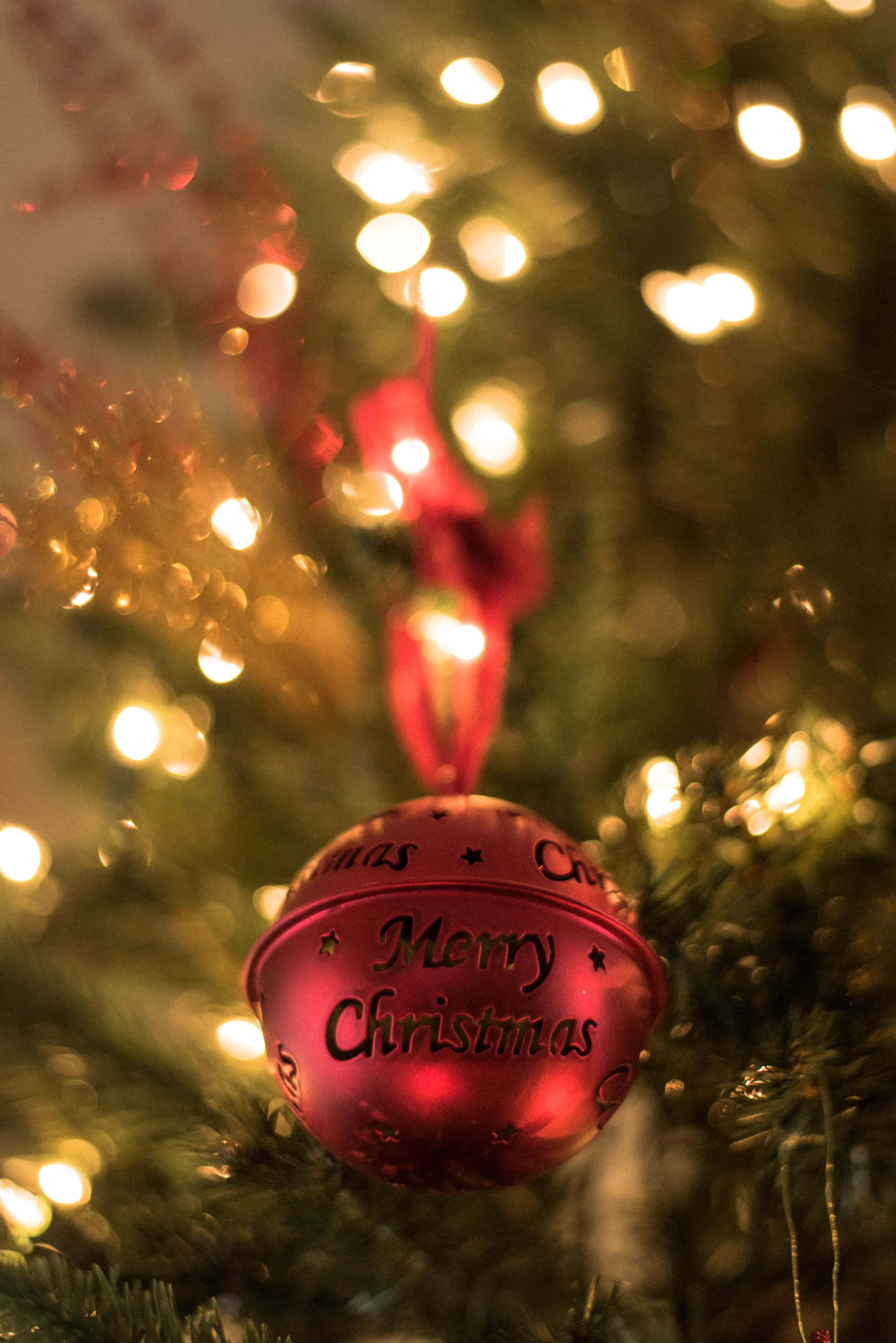 iPhone 聖誕風紅色鈴鐺壁紙 壁紙 — 一個閃亮的紅色聖誕裝飾，上面有「Merry Christmas」字樣，掛在一棵點綴著溫暖燈光的樹上。