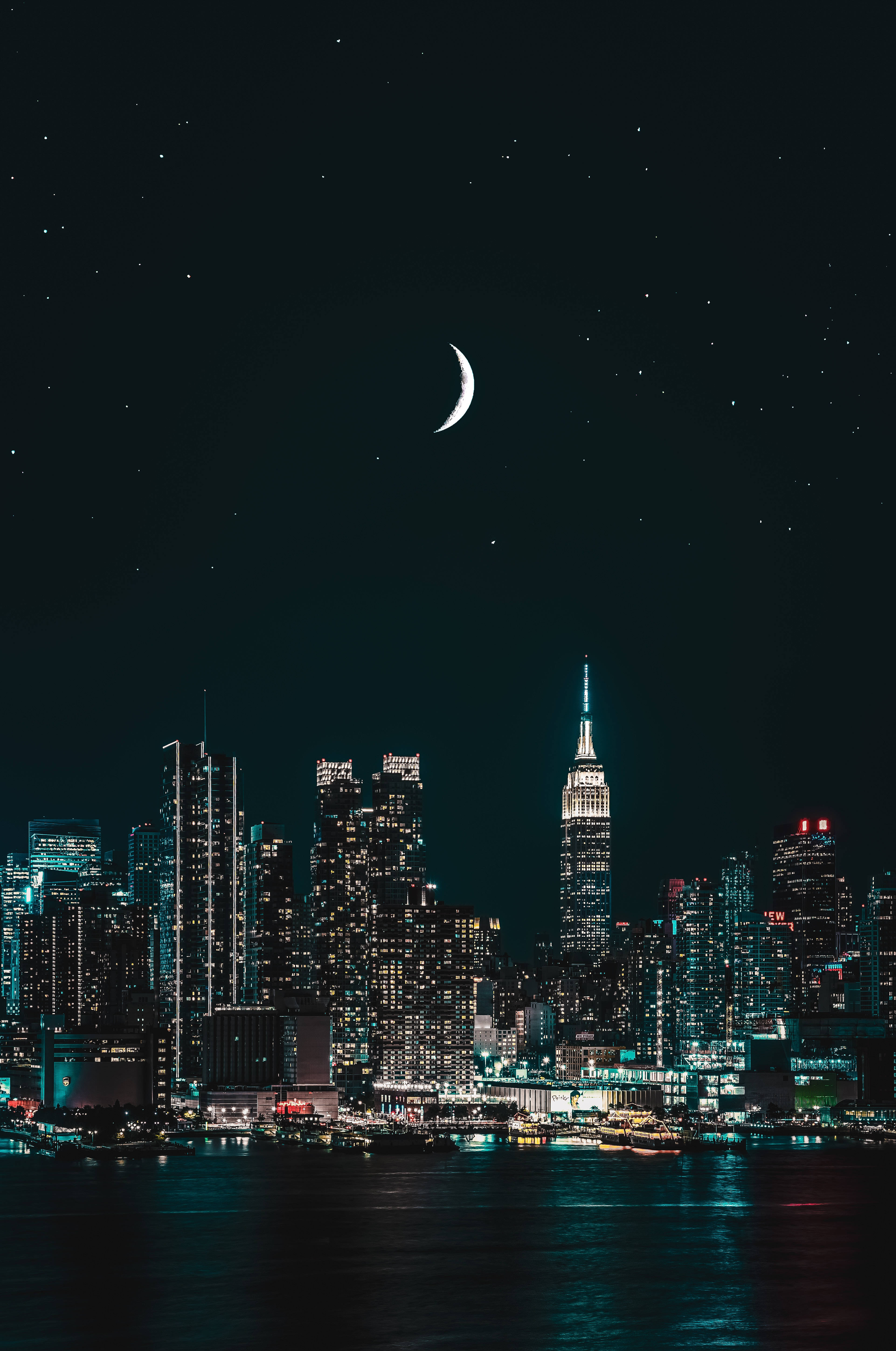 iPhone 解鎖螢幕城市夜景 壁紙 — 一彎新月和星星在夜間照亮紐約市天際線的上方，水面倒映著光芒。