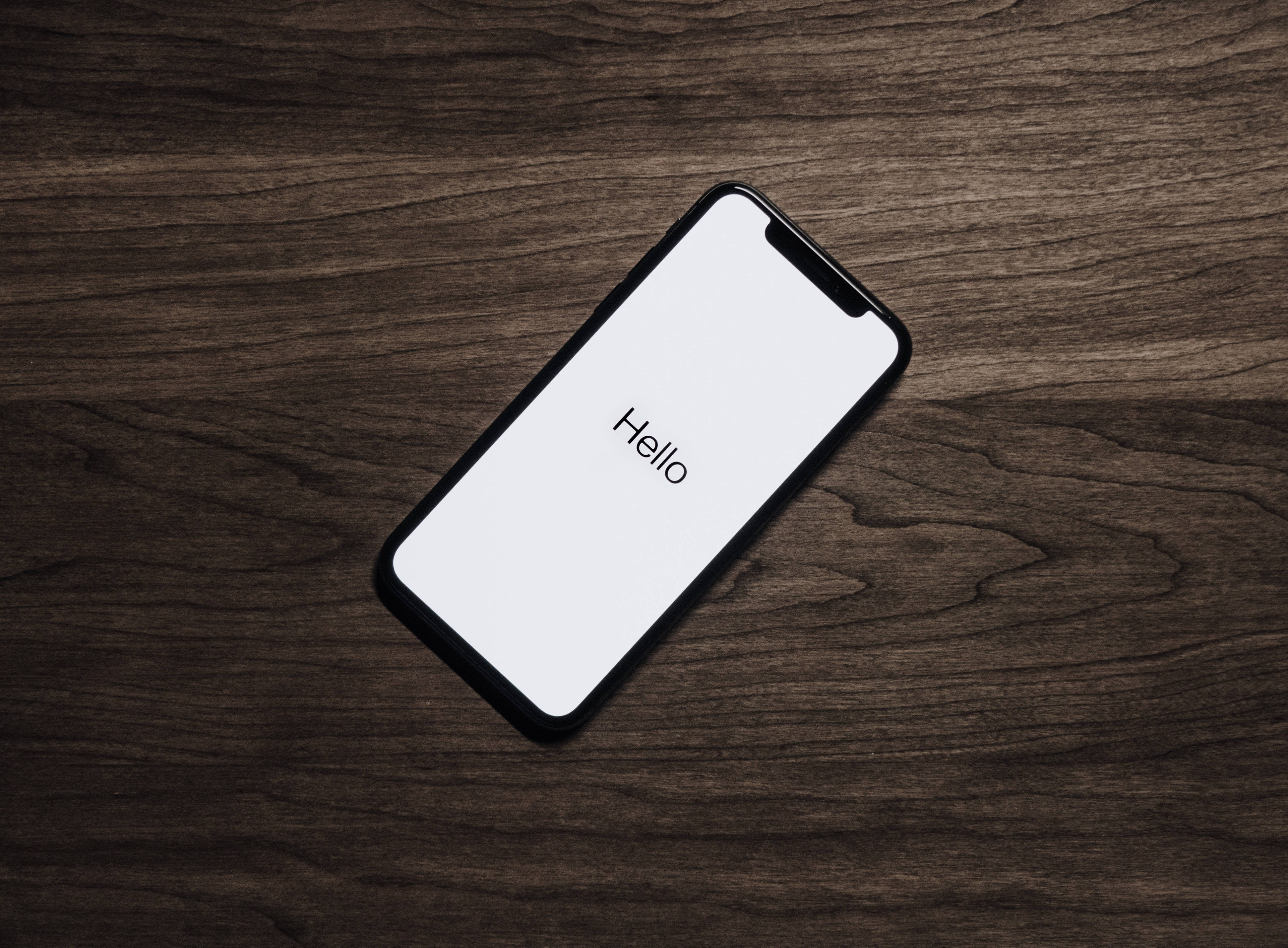 酷iPhone顯示「Hello」 壁紙 — 一部時尚智能手機，白色屏幕上顯示「hello」字樣，斜放在深色紋理木質表面上。