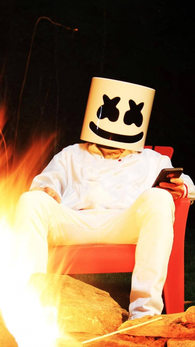 iPhone上的Marshmello DJ藝術圖像 壁紙 — 一個戴著白色棉花糖頭盔的人晚上坐在紅色椅子上，旁邊是熊熊燃燒的營火。