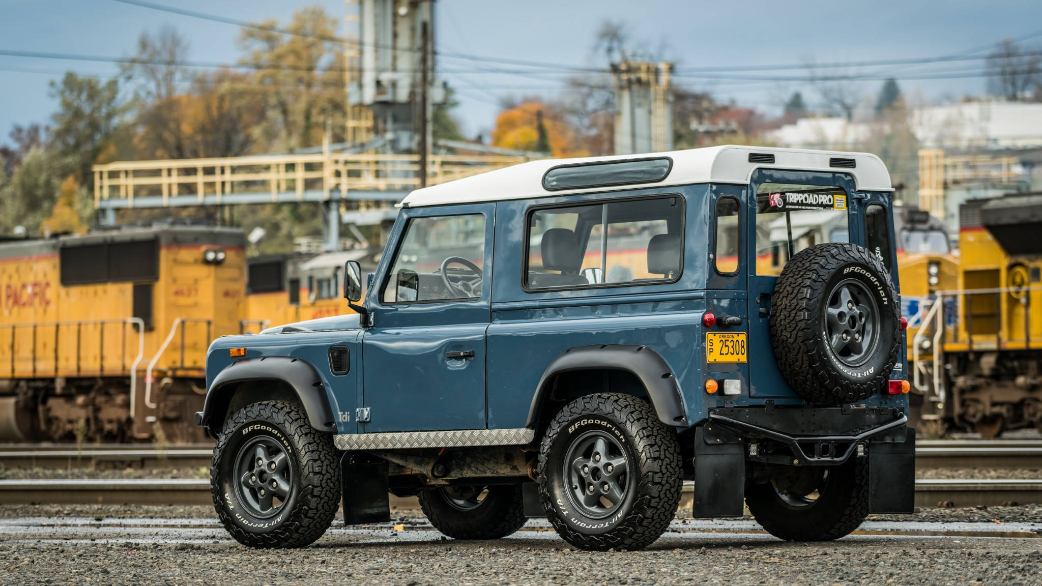 iPhone主題設計：1990年Byron藍色路虎Defender 90 壁紙 — 一輛藍色 Land Rover Defender 堅固地停在鐵軌旁的礫石上，背景有黃色貨運車廂和晴朗的天空。