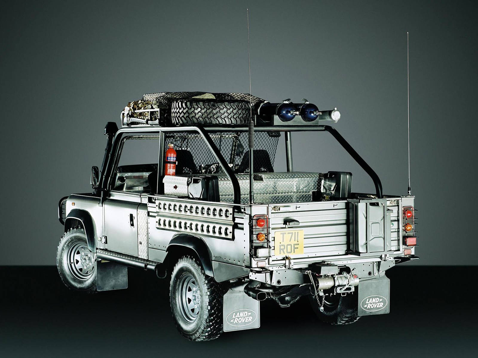 iPhone主題設計：Kyosho Defender 路虎車模 壁紙 — 一輛銀色 Land Rover Defender 皮卡車，配有車頂行李架、備用輪胎和緊急燈，停在深灰色工作室背景前。