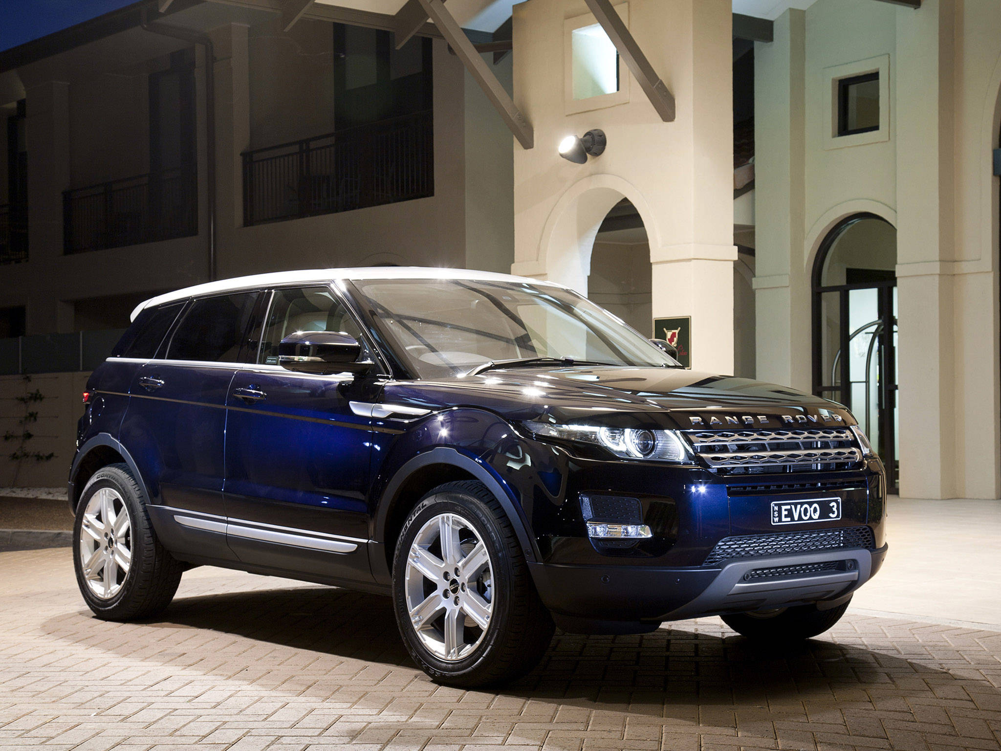 iPhone主題設計：路虎Range Evoque藍色車款 壁紙 — 一輛時尚的深藍色Range Rover Evoque在夜間停在一棟帶有拱形門廊的現代建築前。