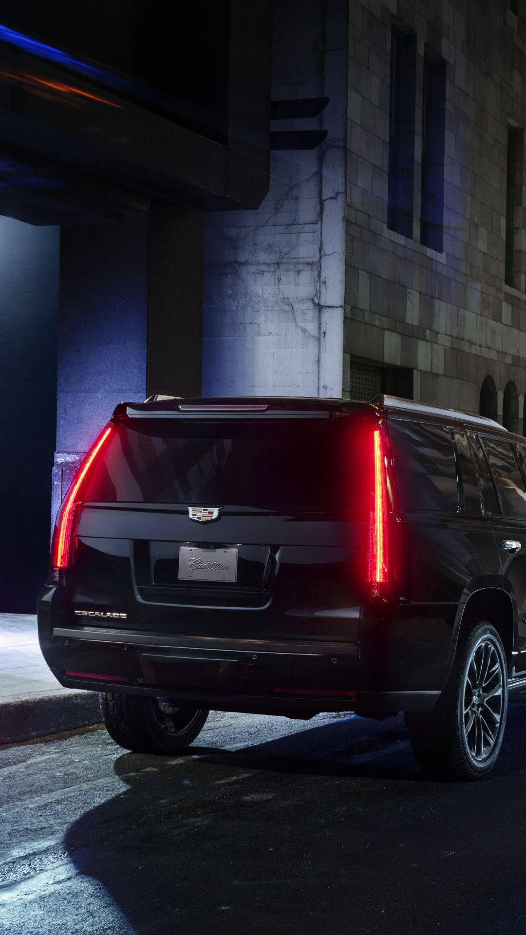 iPhone拍攝的夜間街頭黑色凱迪拉克 壁紙 — 一輛黑色 Cadillac Escalade SUV 在夜間停在一座石建築前的橋下，散發出鮮豔的紅色尾燈光芒。