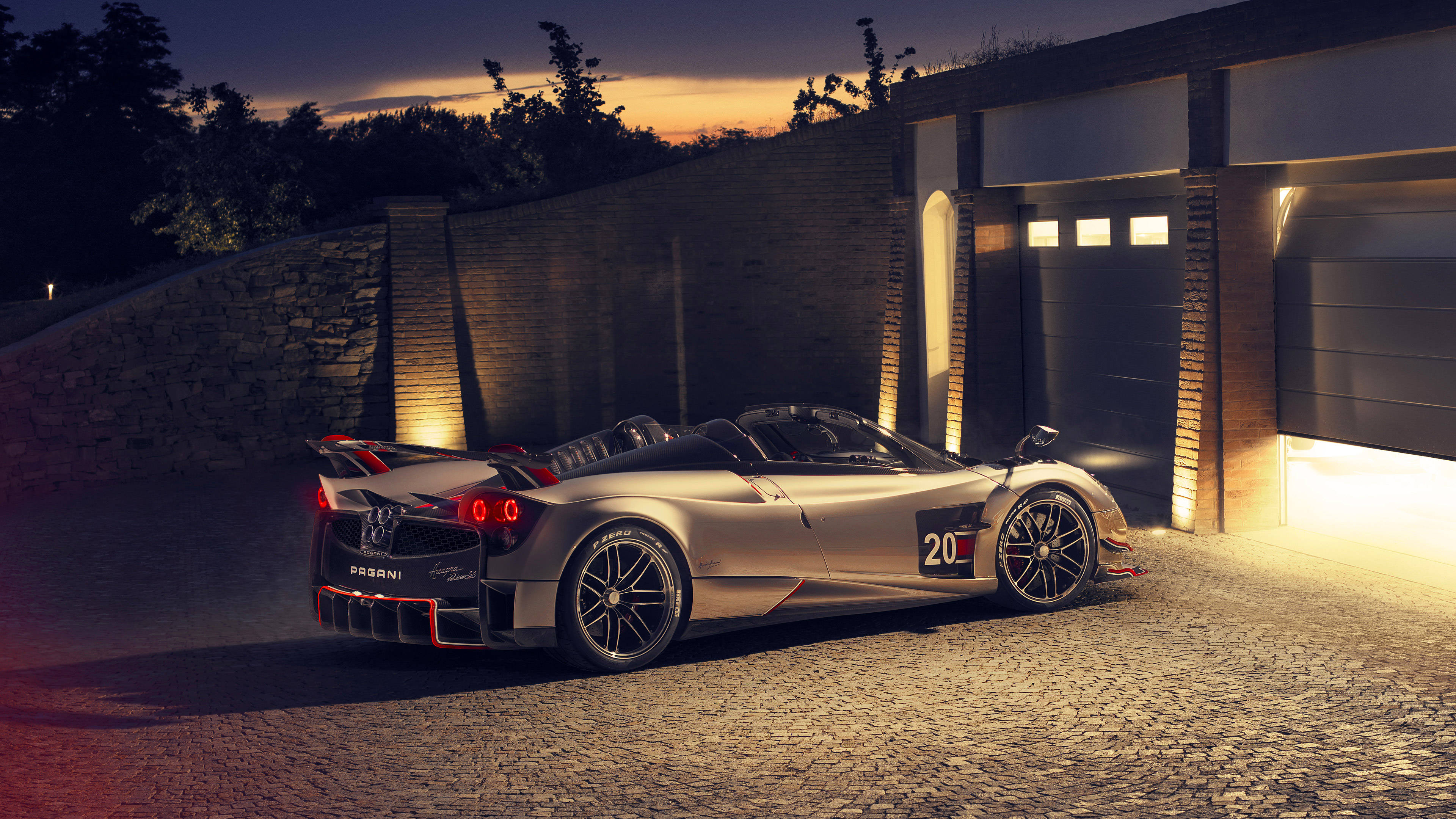 iPhone拍攝的帕加尼Huayra Roadster BC超跑 壁紙 — 一輛銀色Pagani Huayra Roadster在暮色燈光下閃閃發光，停在現代車庫旁的鵝卵石車道上。