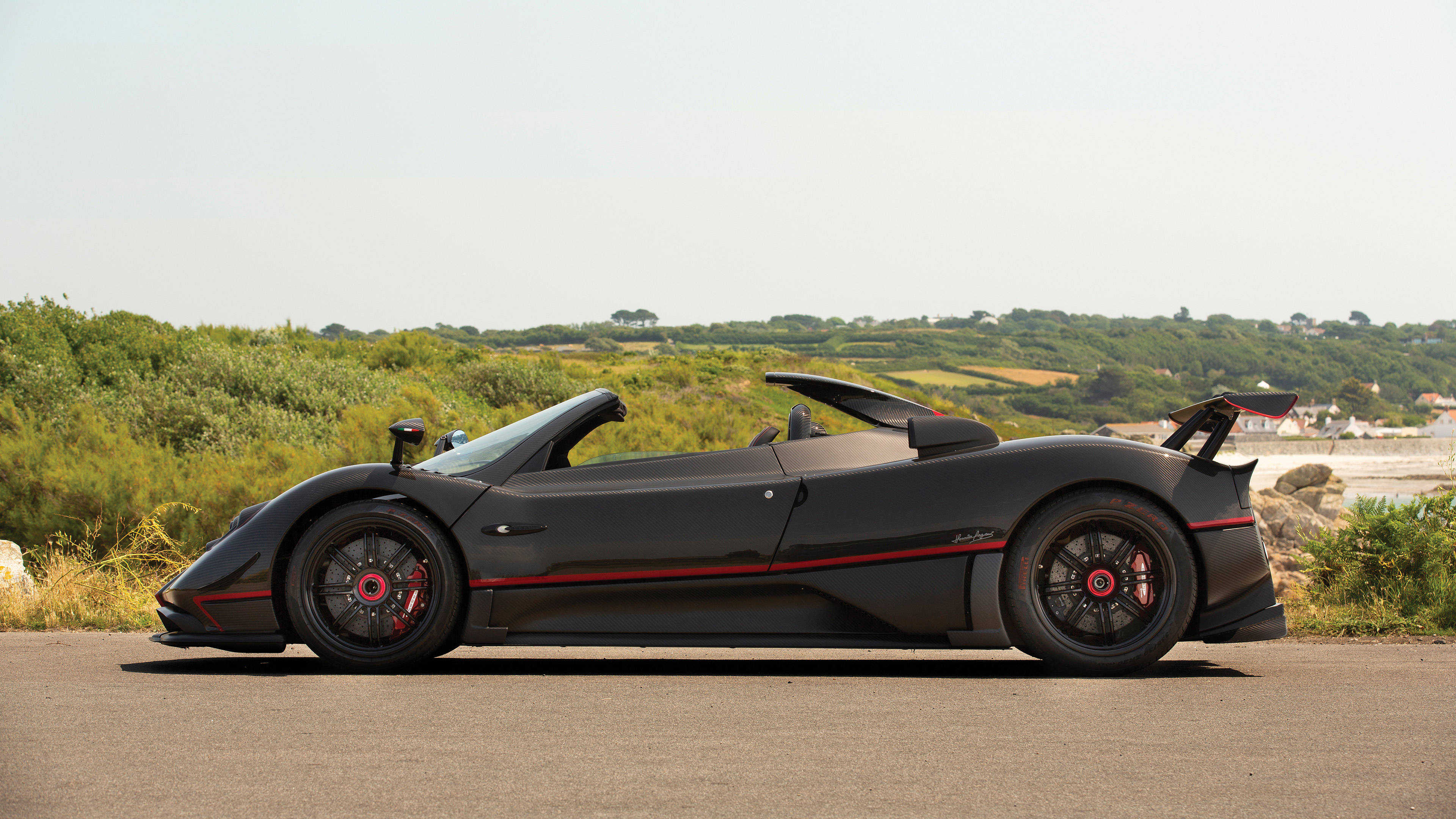 iPhone拍攝的帕加尼Zonda Aether側視圖 壁紙 — 一輛時尚的黑色Pagani Zonda Cinque Roadster，帶有紅色點綴，停泊在明亮天空下綠丘旁的沿海道路上。