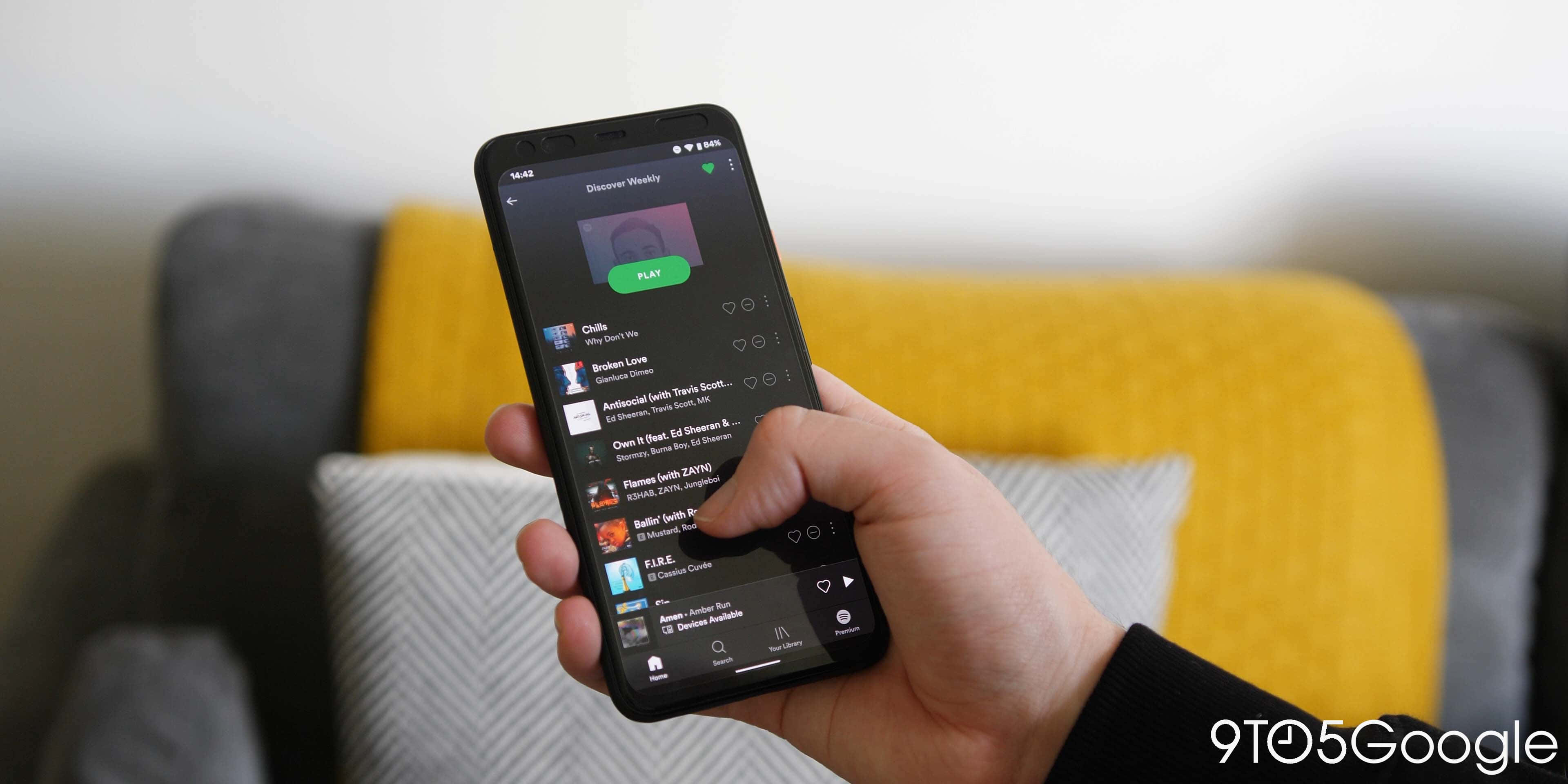iPhone播放Spotify歌曲圖片 壁紙 — 一隻手拿著智能手機，顯示音樂應用程式的播放列表，背景是帶有黃色和灰色靠墊的舒適沙發。