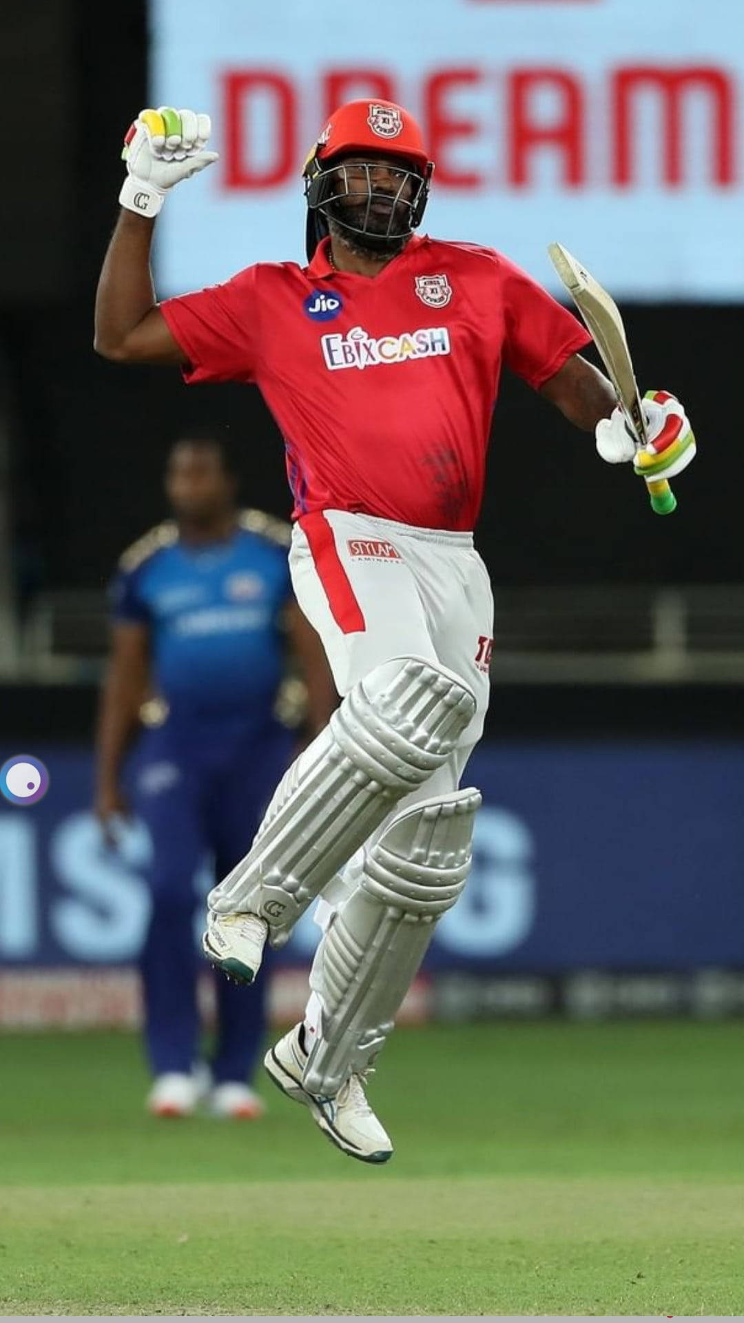 IPL 2021 克里斯·蓋勒握拳慶祝 壁紙 — 一名身穿紅色球衣的板球運動員手持球棒和手套，在綠色球場上歡樂地跳起慶祝勝利。