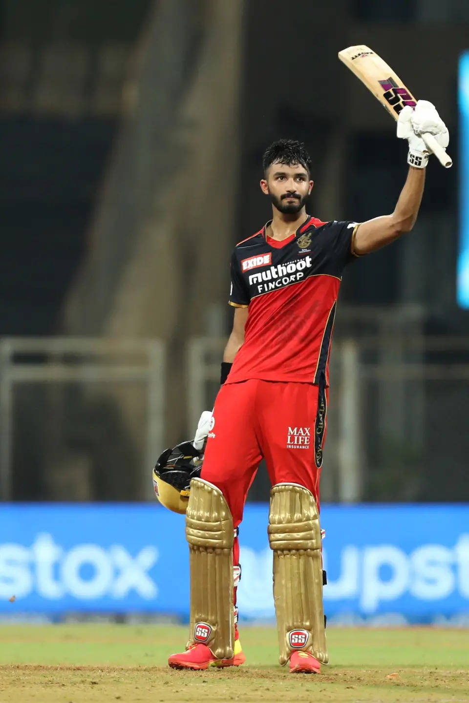 IPL 2021 德夫杜特·帕迪卡尔 高舉球棒 壁紙 — 身穿紅色球衣的板球運動員在明亮的燈光下於球場上勝利地高舉球棒。