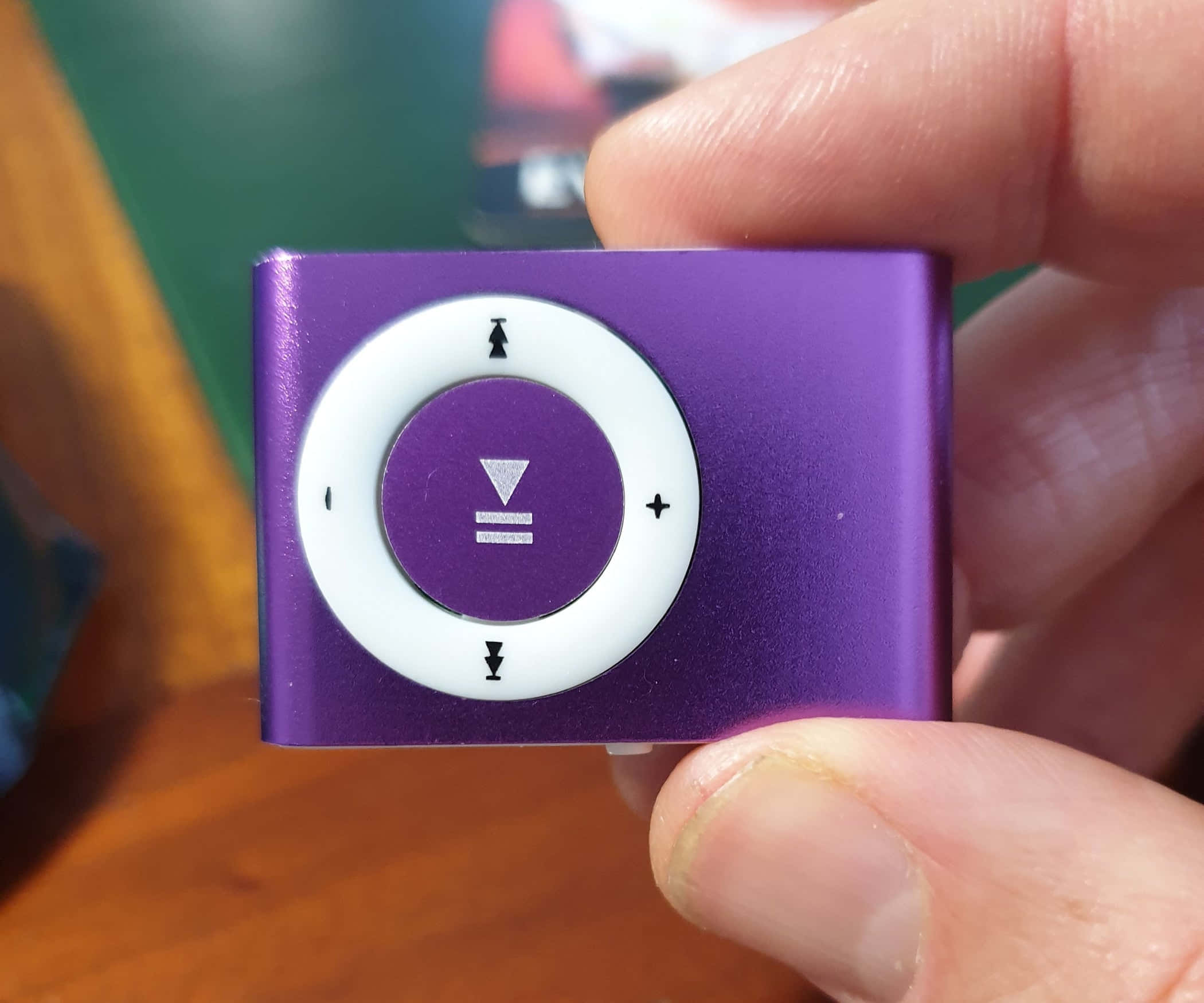 iPod Shuffle MP3圖片 壁紙 — 一隻手拿著一個帶有白色點擊輪的紫色 iPod shuffle，背景是模糊的木紋。